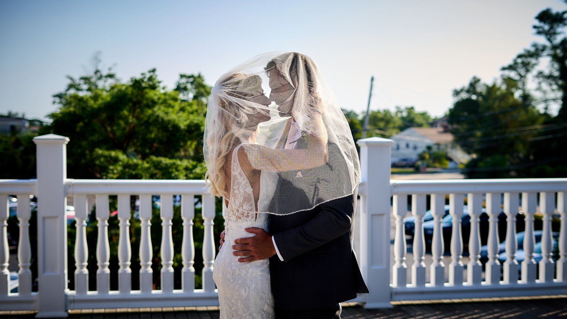 A bride and groom are kissing under a veil on a balcony.