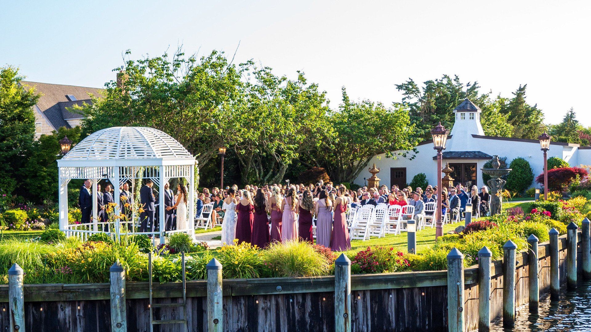 A large group of people are gathered in front of a gazebo for a wedding ceremony.