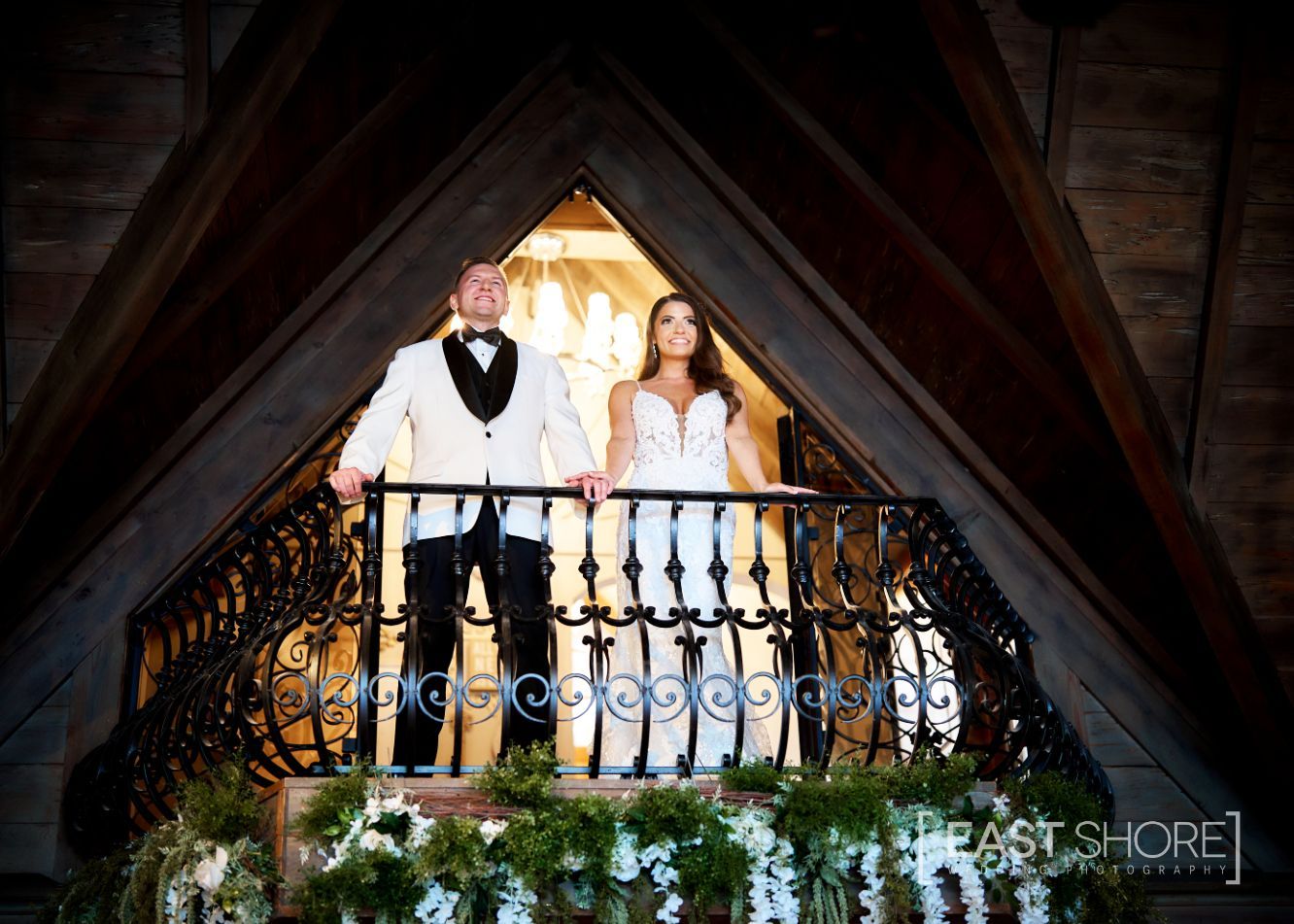 A bride and groom are standing on a balcony holding hands.