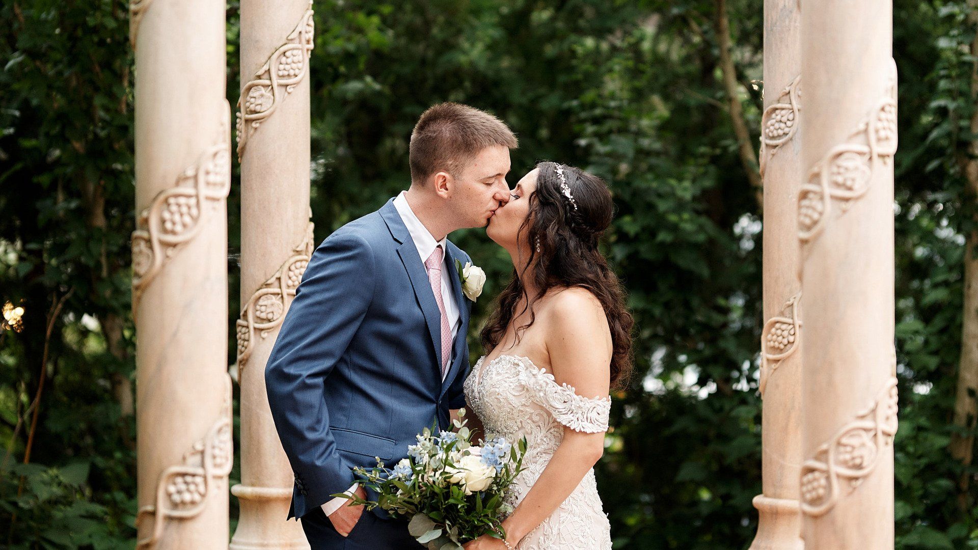 A bride and groom are kissing in front of a row of columns.