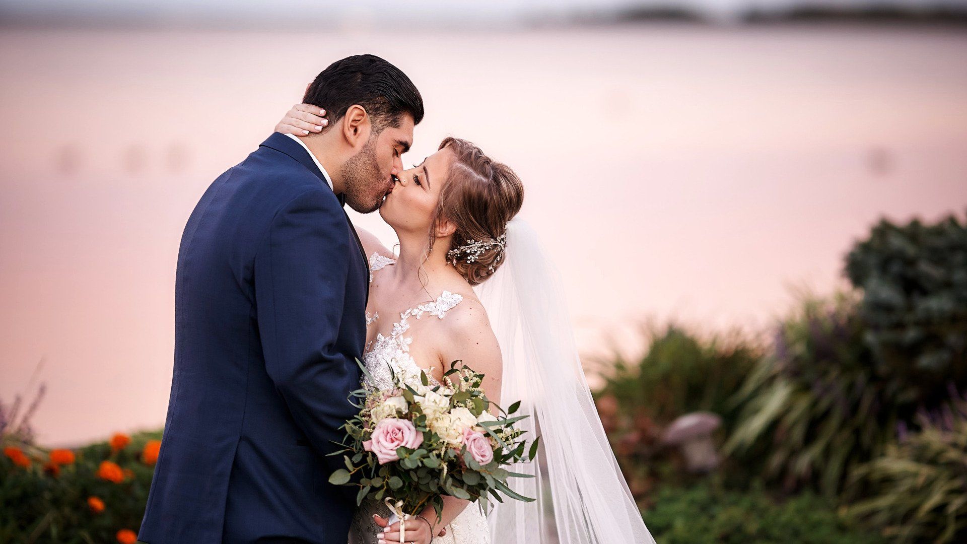 A bride and groom are kissing in front of a body of water.