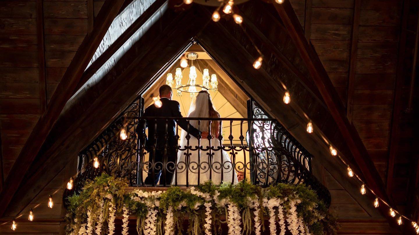 A bride and groom are standing on a balcony holding hands.