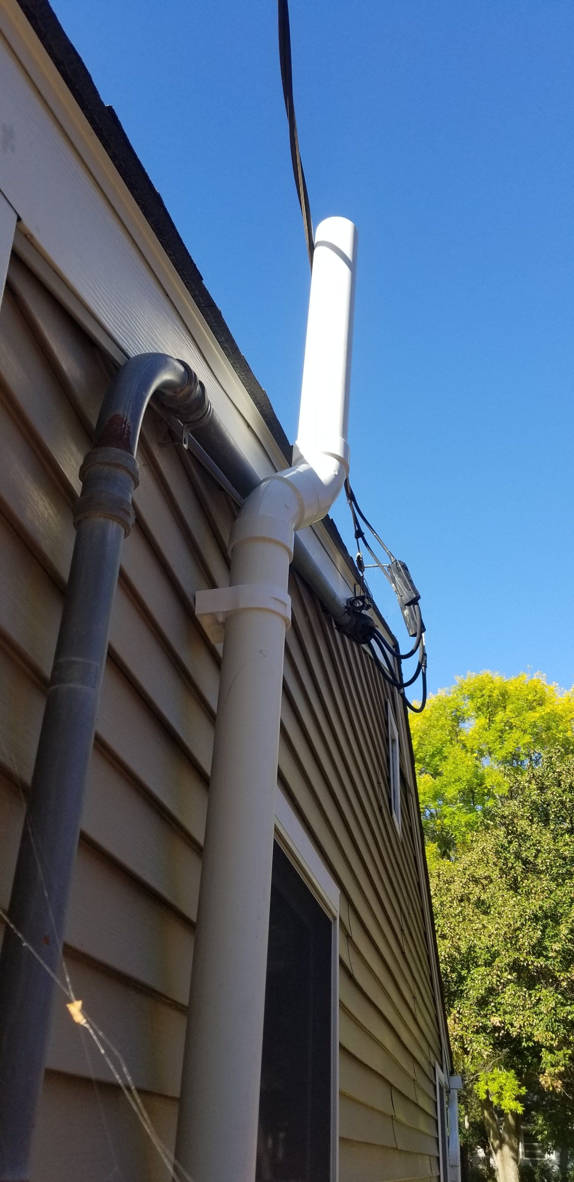 A white PVC pipe running vertically up the side of a building, next to a black pipe, against a blue sky.