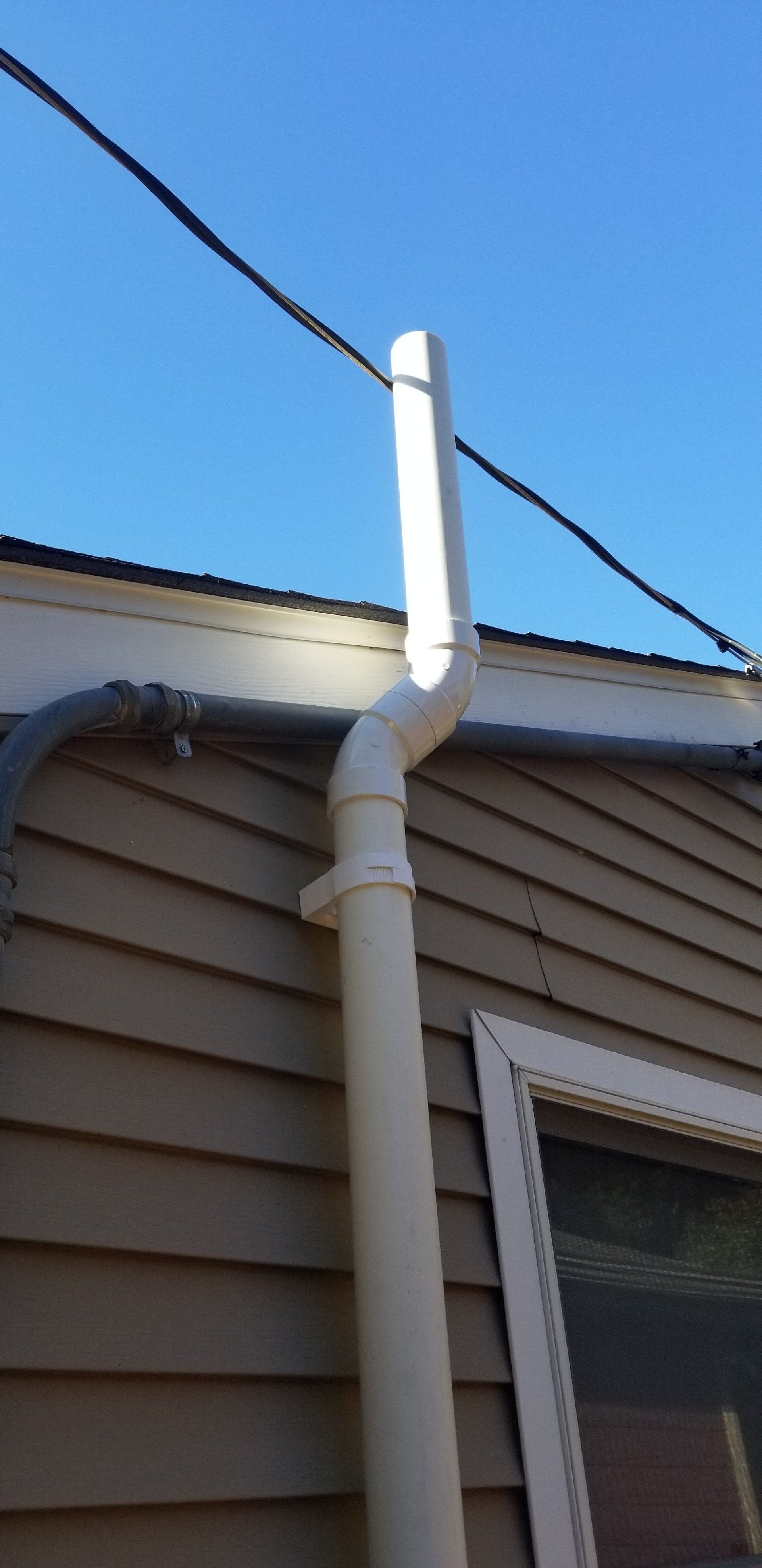 White plastic gutter extending upwards along a beige-sided building, blue sky backdrop.