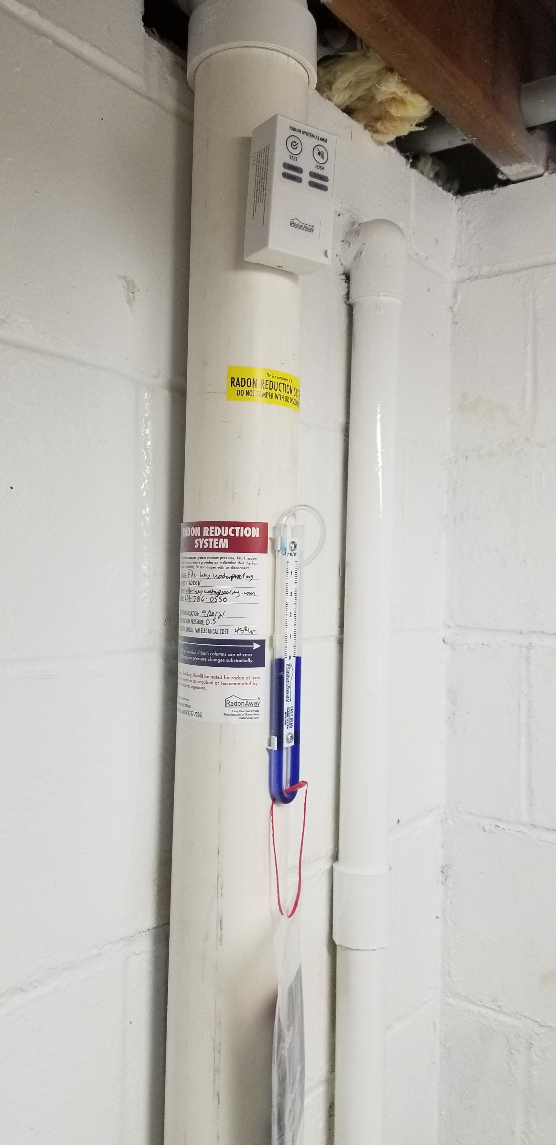 White pipes against a white brick wall. A device is attached to a pipe with labels and a thermometer.