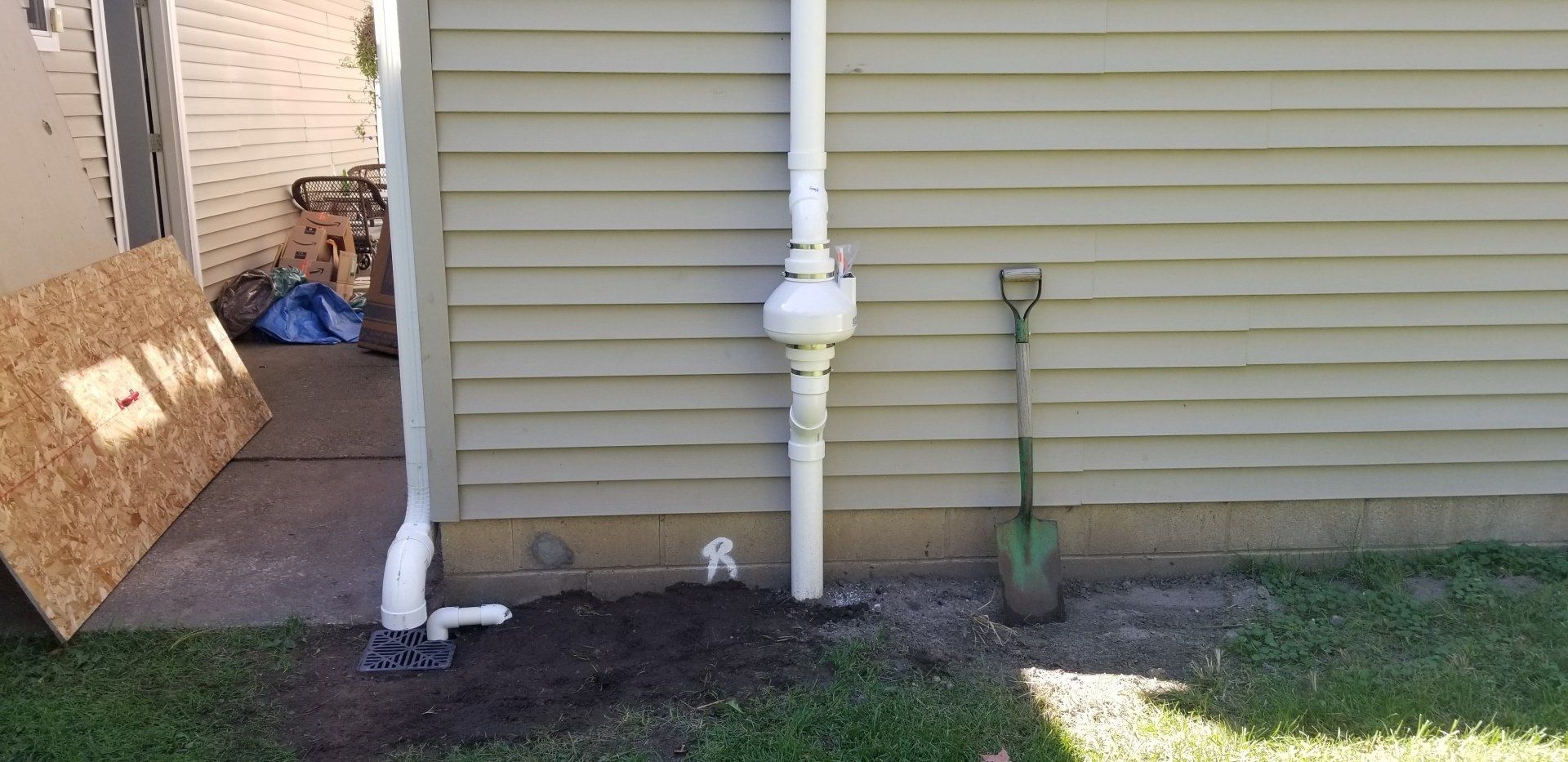 A white gas pipe is on a siding building; a shovel rests in dirt beside it.