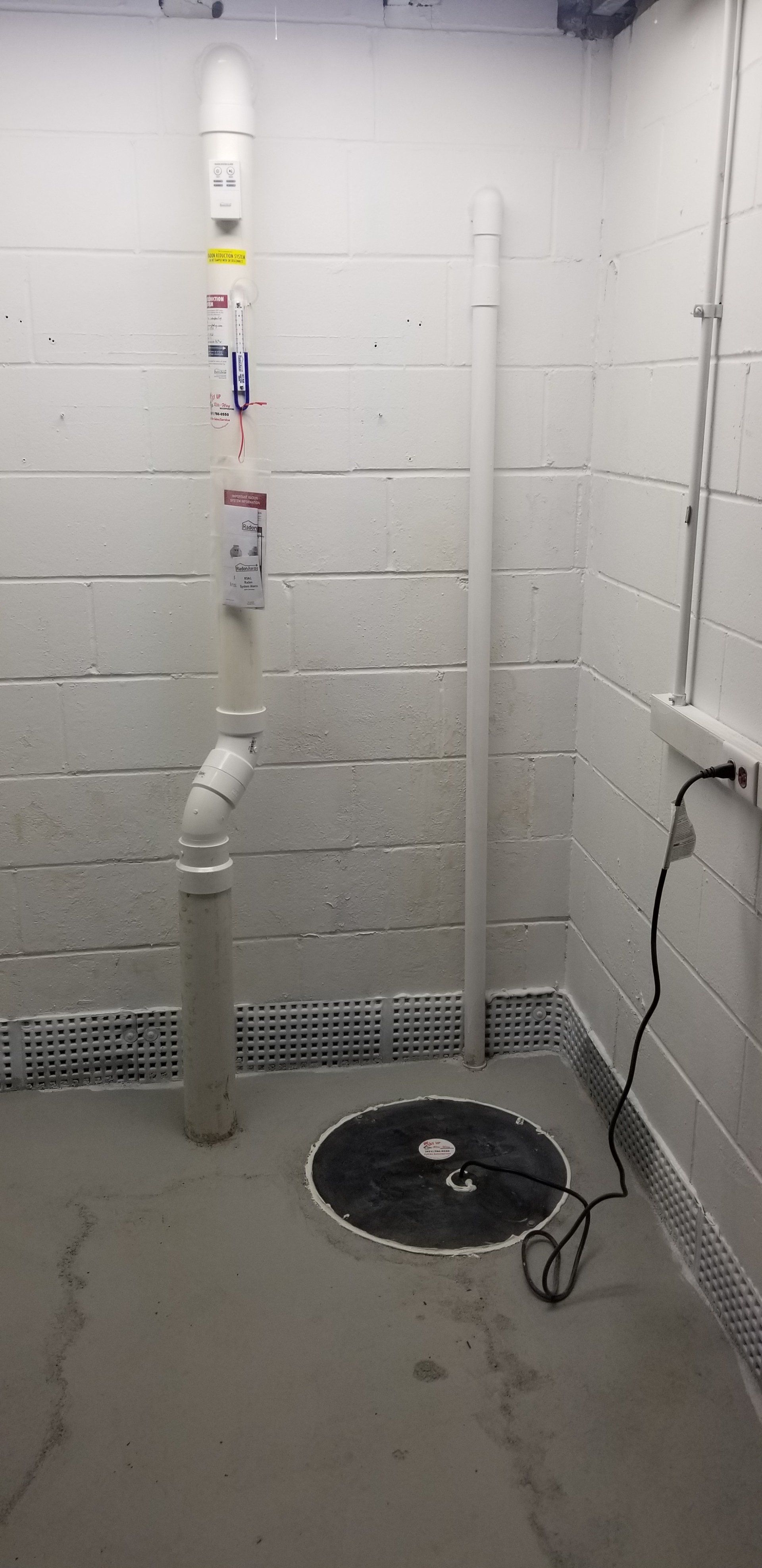 Sump pump system in a concrete-walled room, with a black cover and vertical pipe.