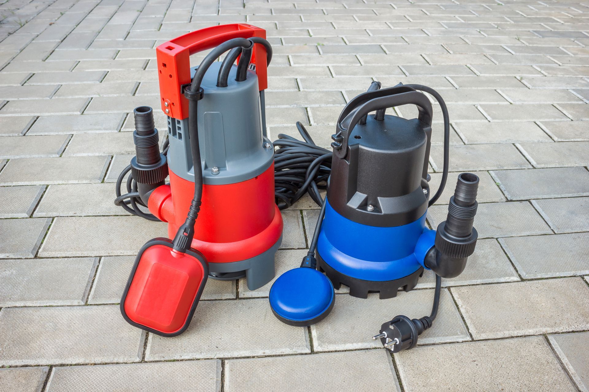 Two submersible sump pumps with hoses and cords placed on a paved surface. Two submersible sump pumps with hoses and cords placed on a paved surface.