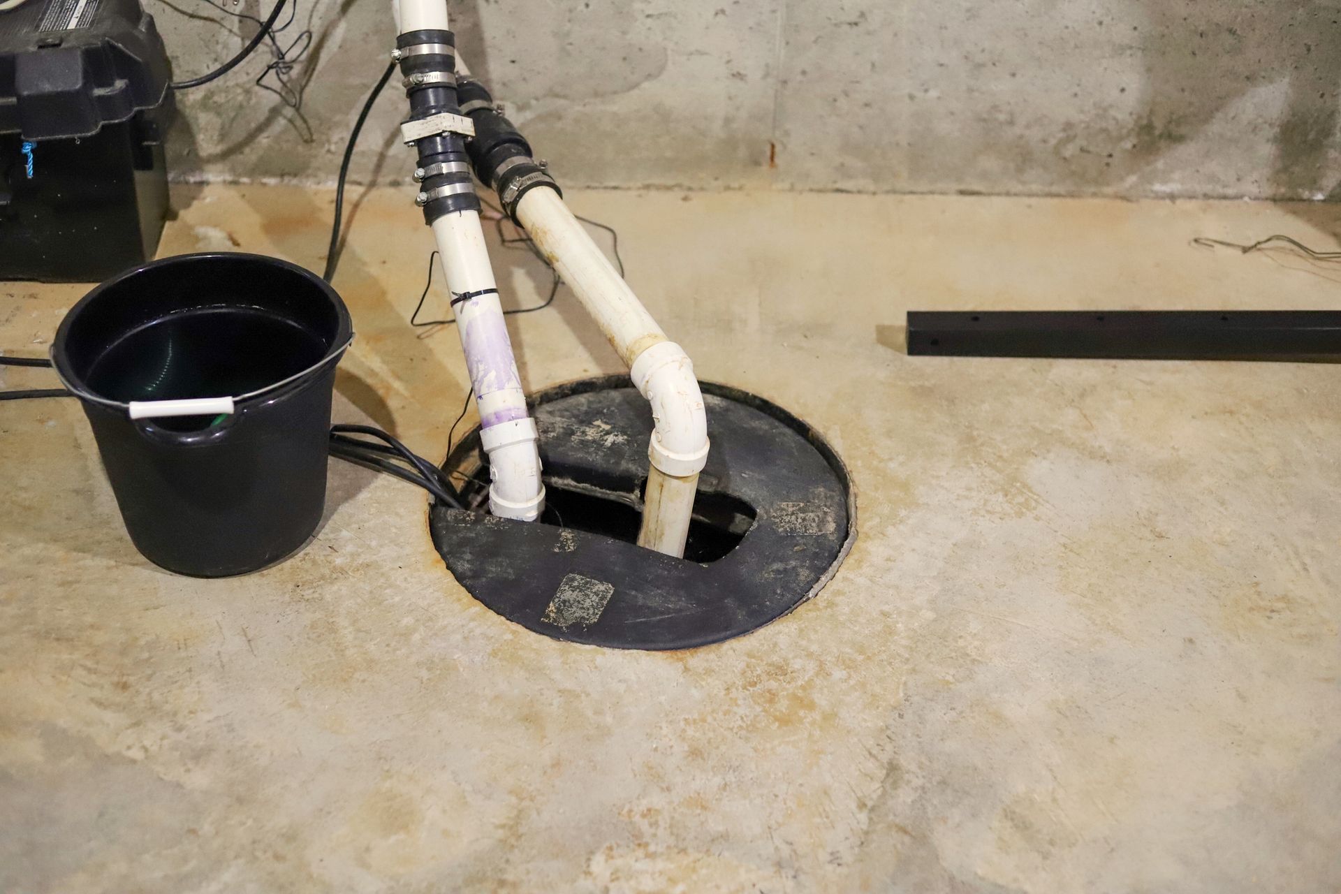 A white PVC sump pump in a basement’s floor, next to a black bucket.