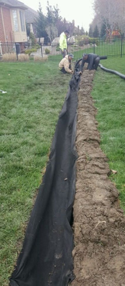 Workers installing a drainage system in a grassy yard, using black fabric in a trench.