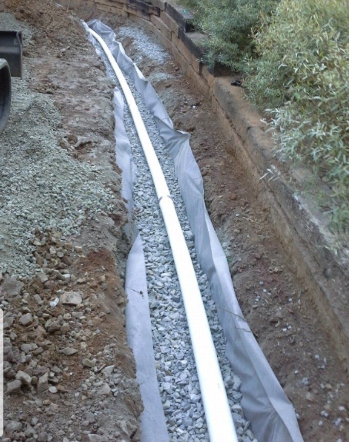Trench with white pipe, gravel, and fabric, likely for drainage, along a hillside.