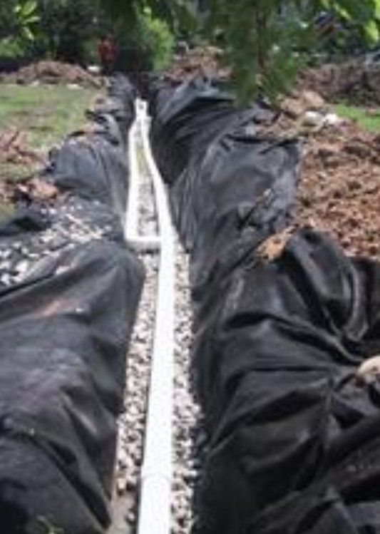 Trench with a white pipe surrounded by gravel, lined with black fabric, in an outdoor setting.