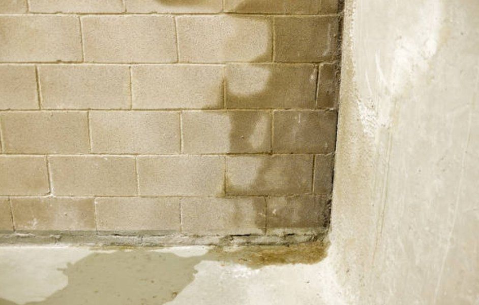 Damp concrete block wall with water stains at the base, water on the floor, and a smooth plastered wall in the corner.