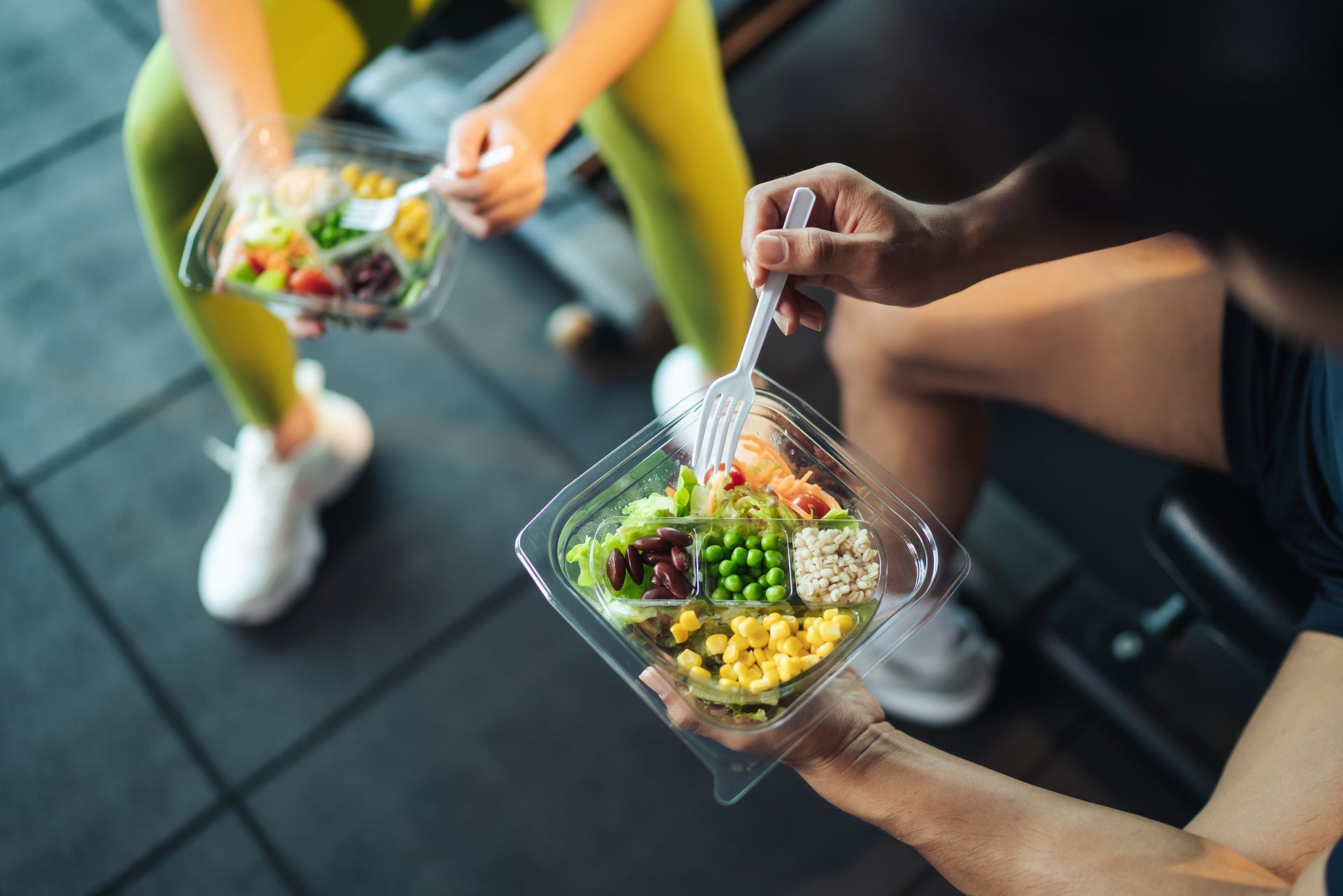 Two people in athletic wear eating salads in a gym.