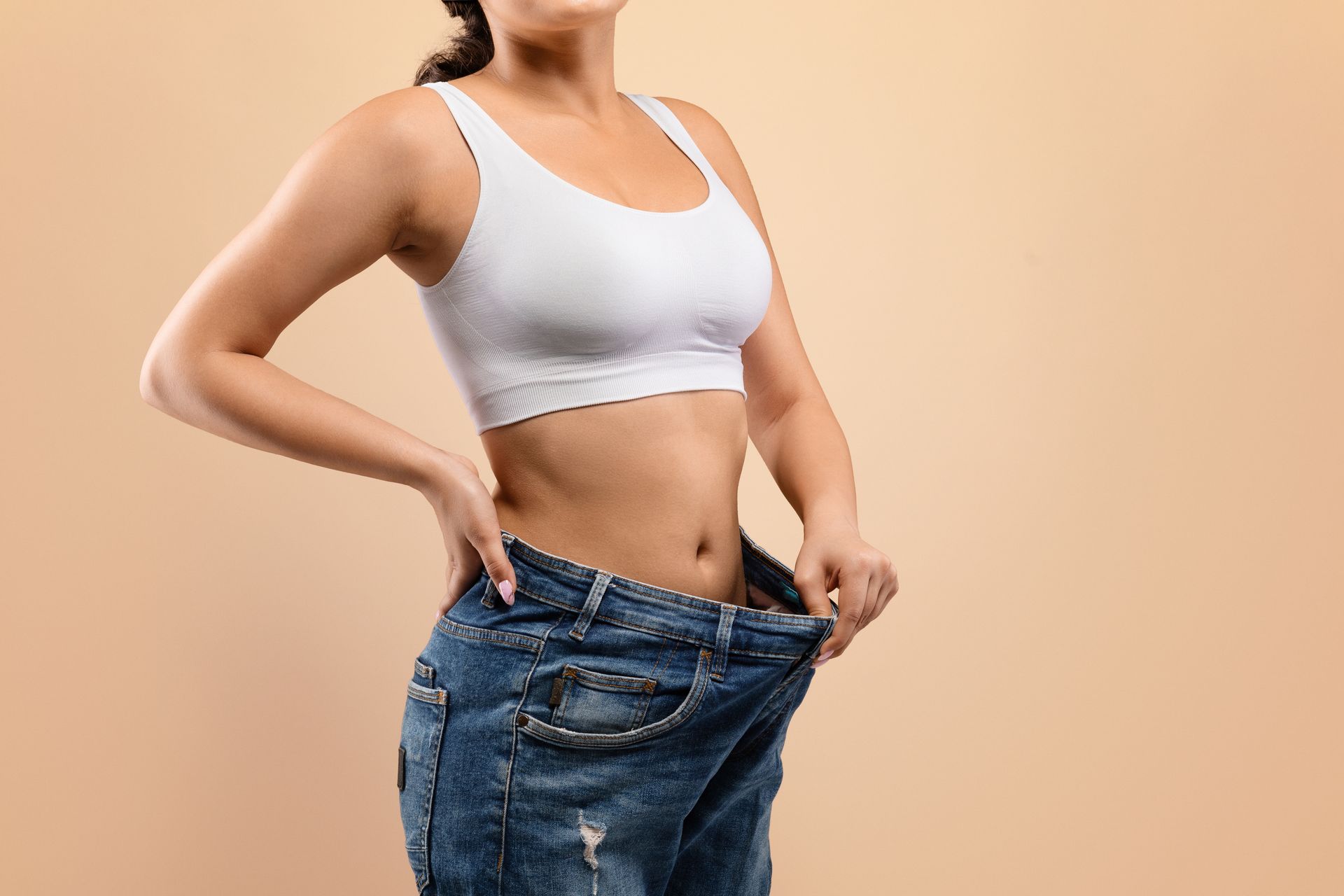 Woman in oversized jeans, hands on waist, showcasing weight loss, wearing white sports bra, tan background.
