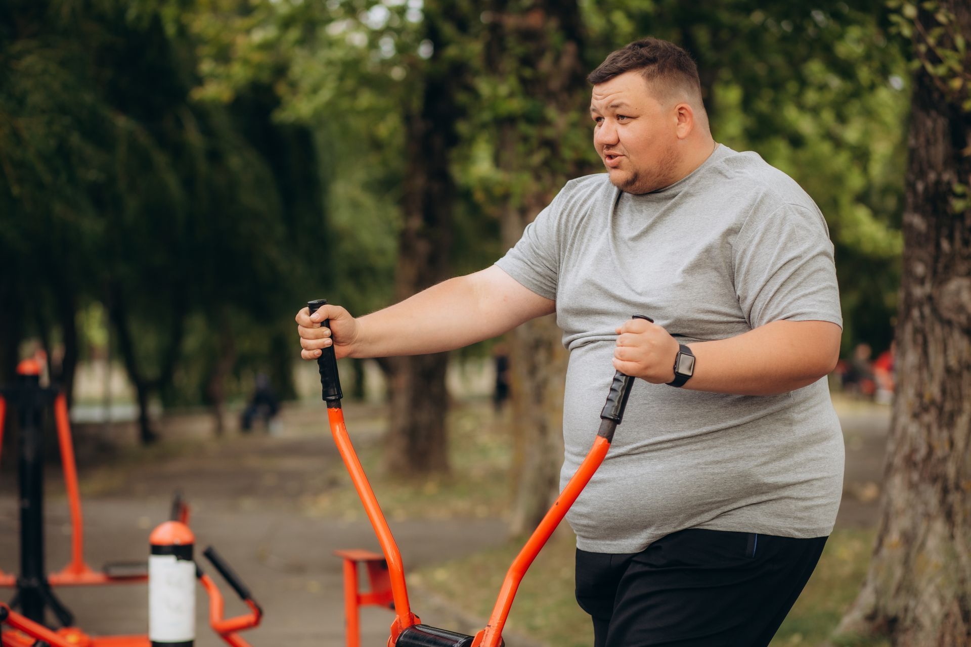 A person uses an outdoor elliptical machine in a park, wearing a grey shirt and black shorts.