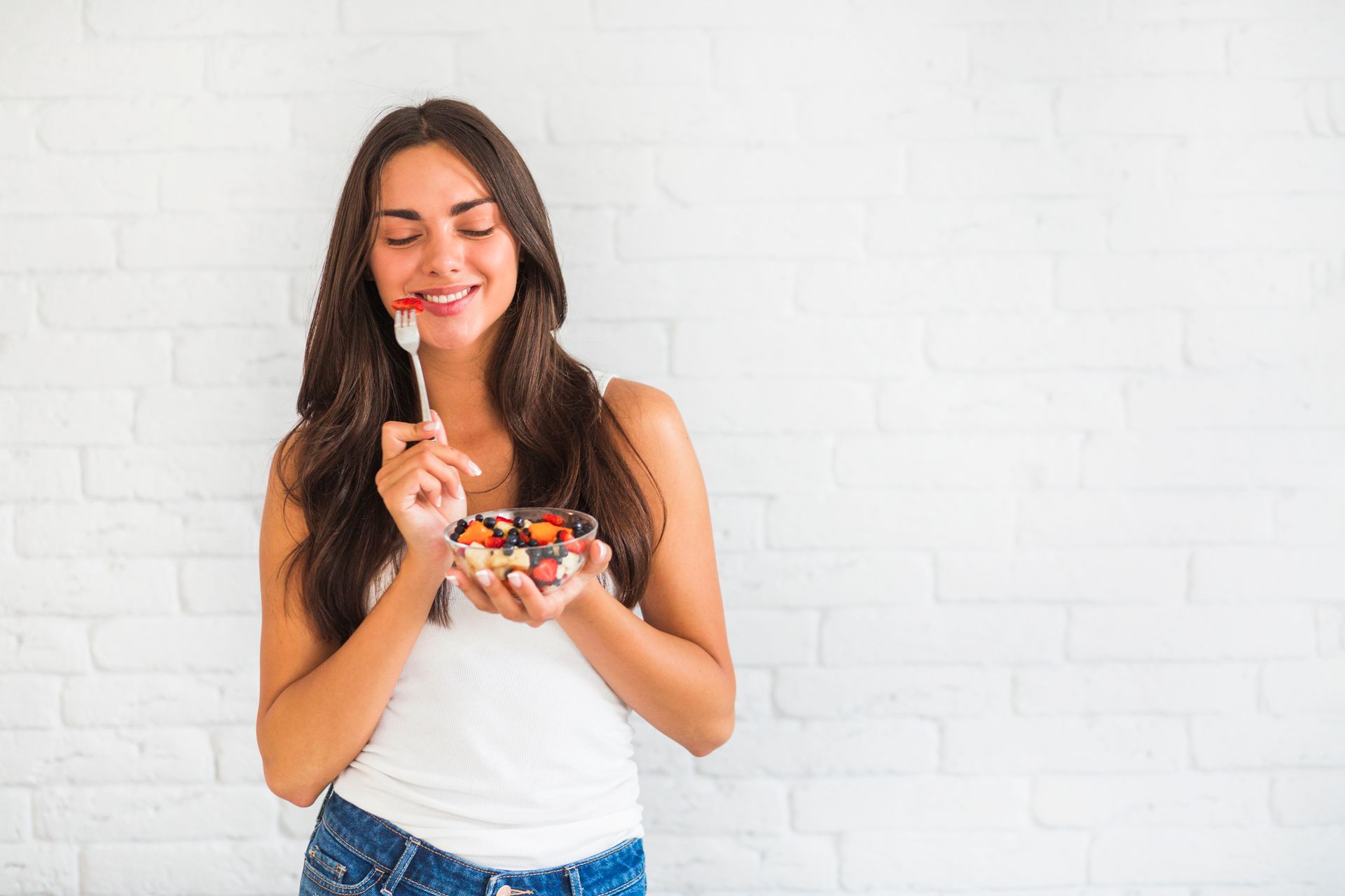 happy young woman standing against white wall eating fruit salad