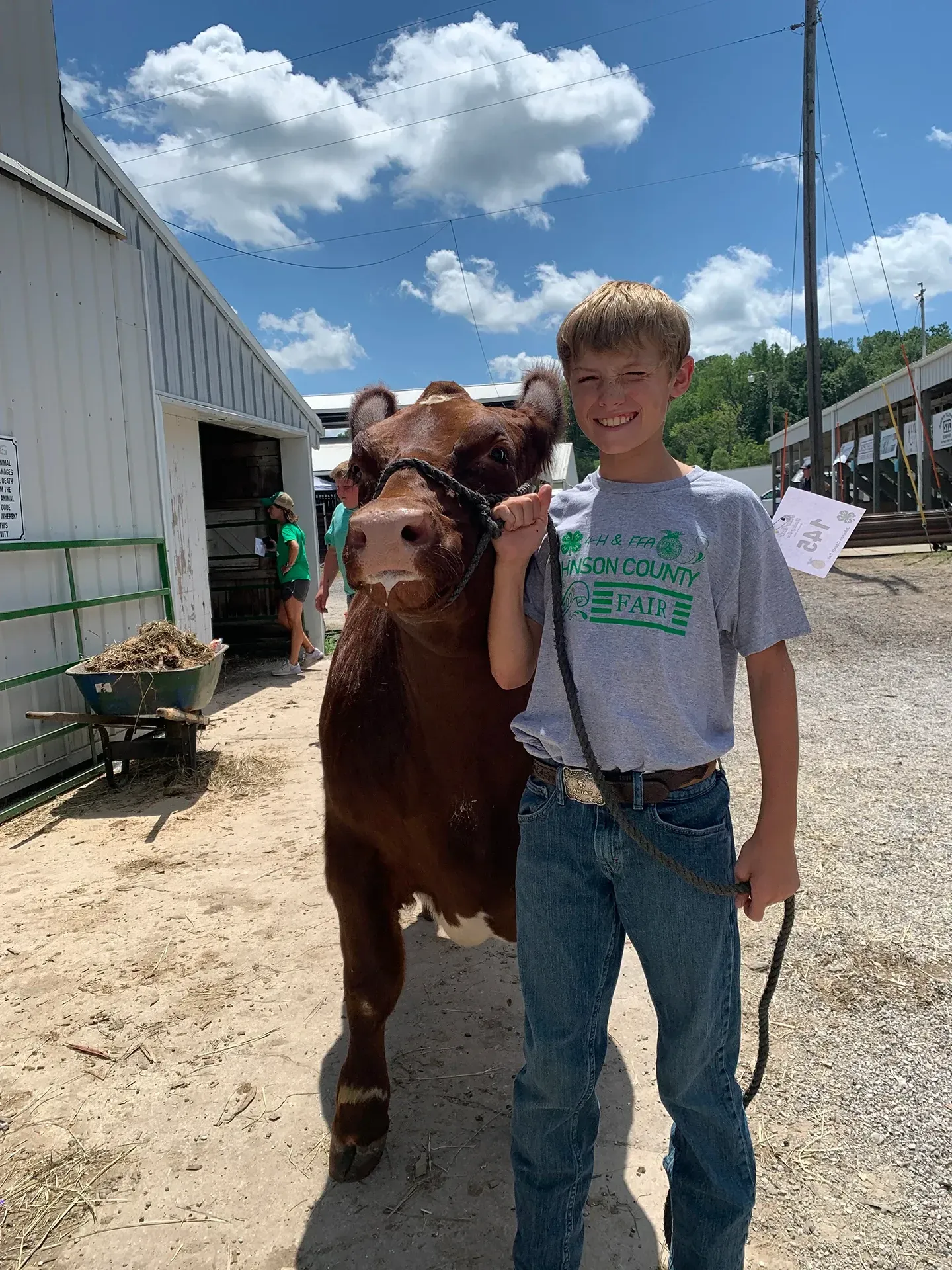 Boy standing beside a brown calf on a sunny farm, with barns and a blue sky in the background