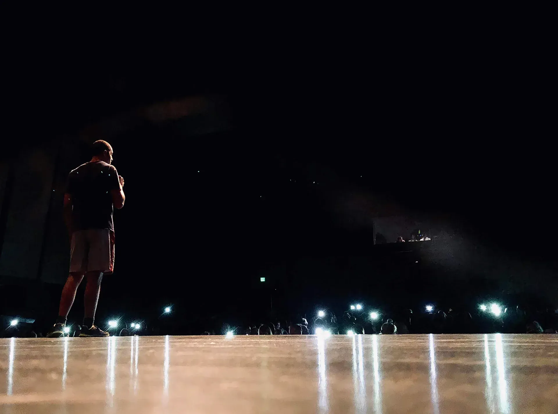 Person stands on a stage facing a dark crowd with bright phone lights in the audience.