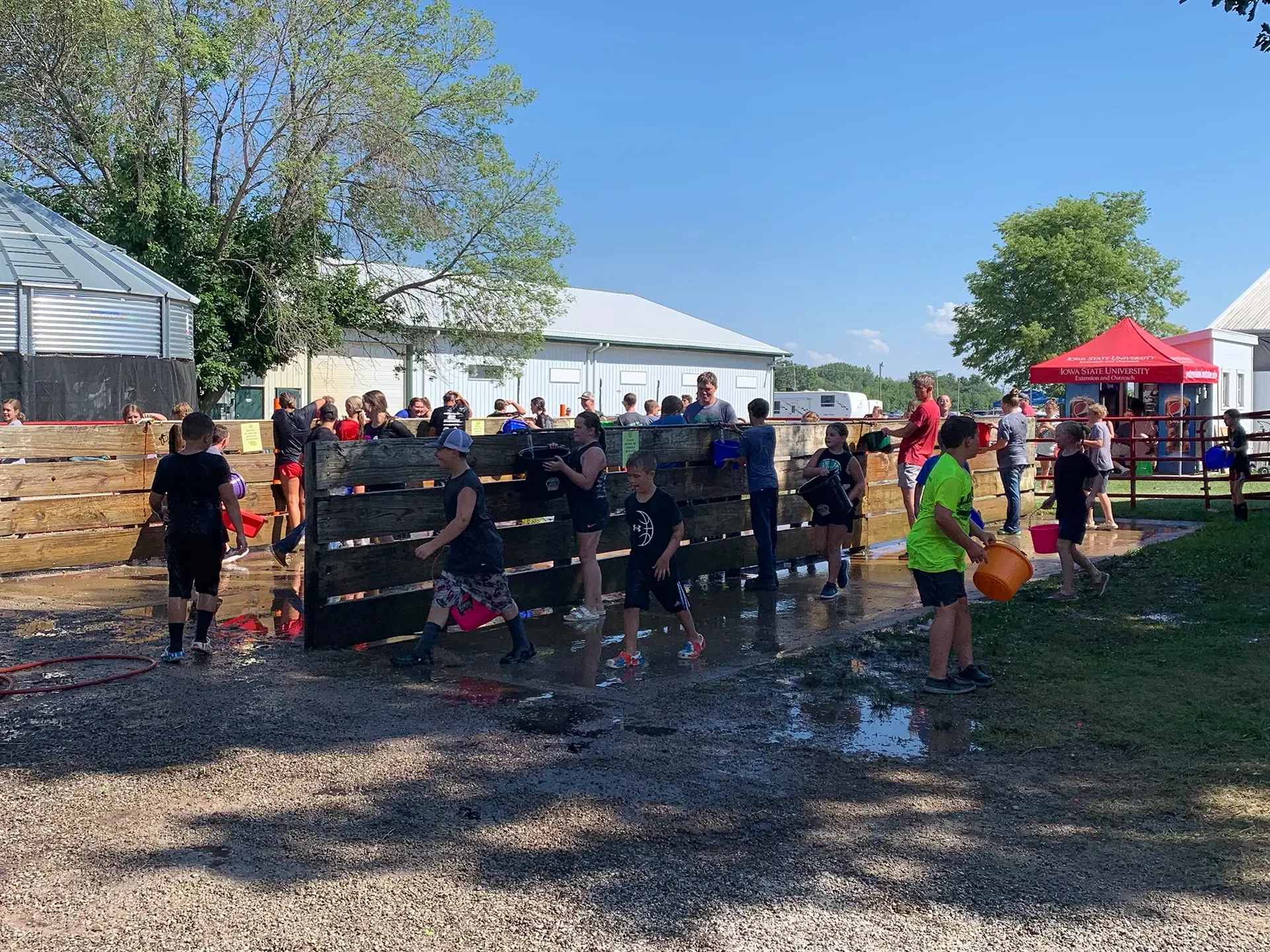 People climbing a muddy obstacle wall at an outdoor event with tents and spectators nearby