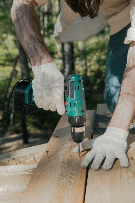 Person wearing gloves using a green power drill to screw into a wooden plank outdoors.