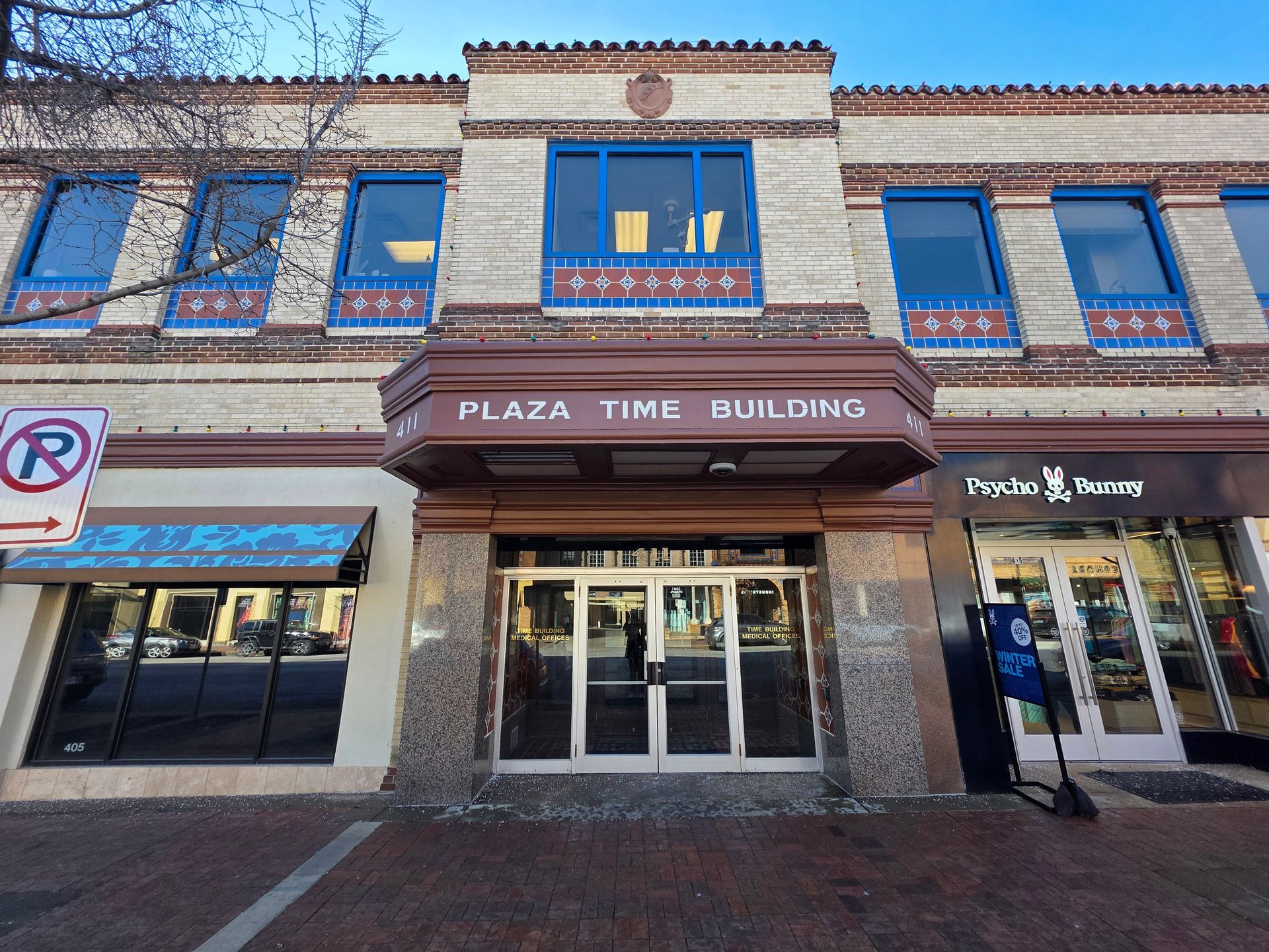 Plaza Time Building facade with brown awning, glass doors, and blue accents on a brick building with storefronts.