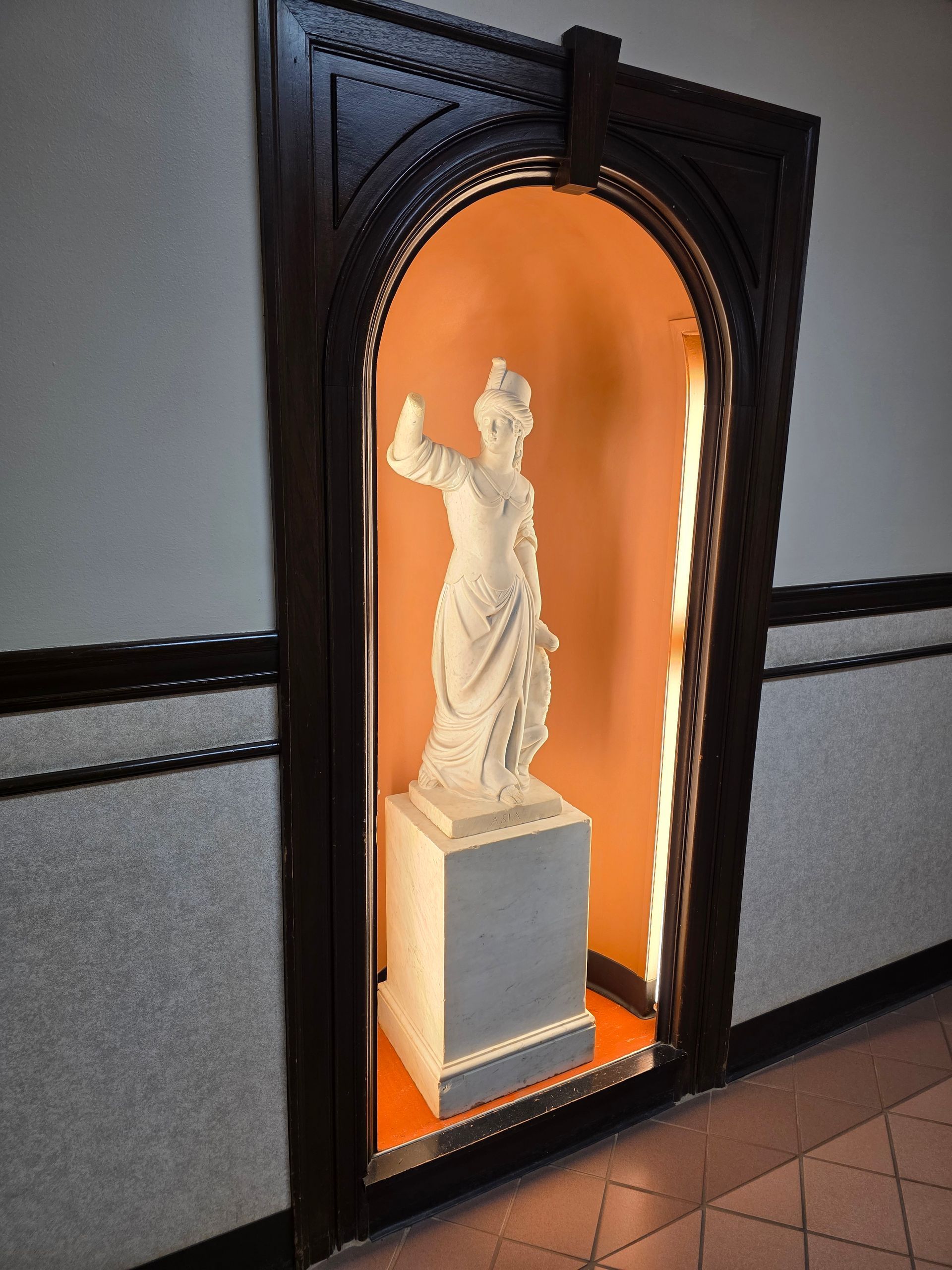 Statue in a dark wooden alcove with orange backlighting. A white statue stands on a gray pedestal.