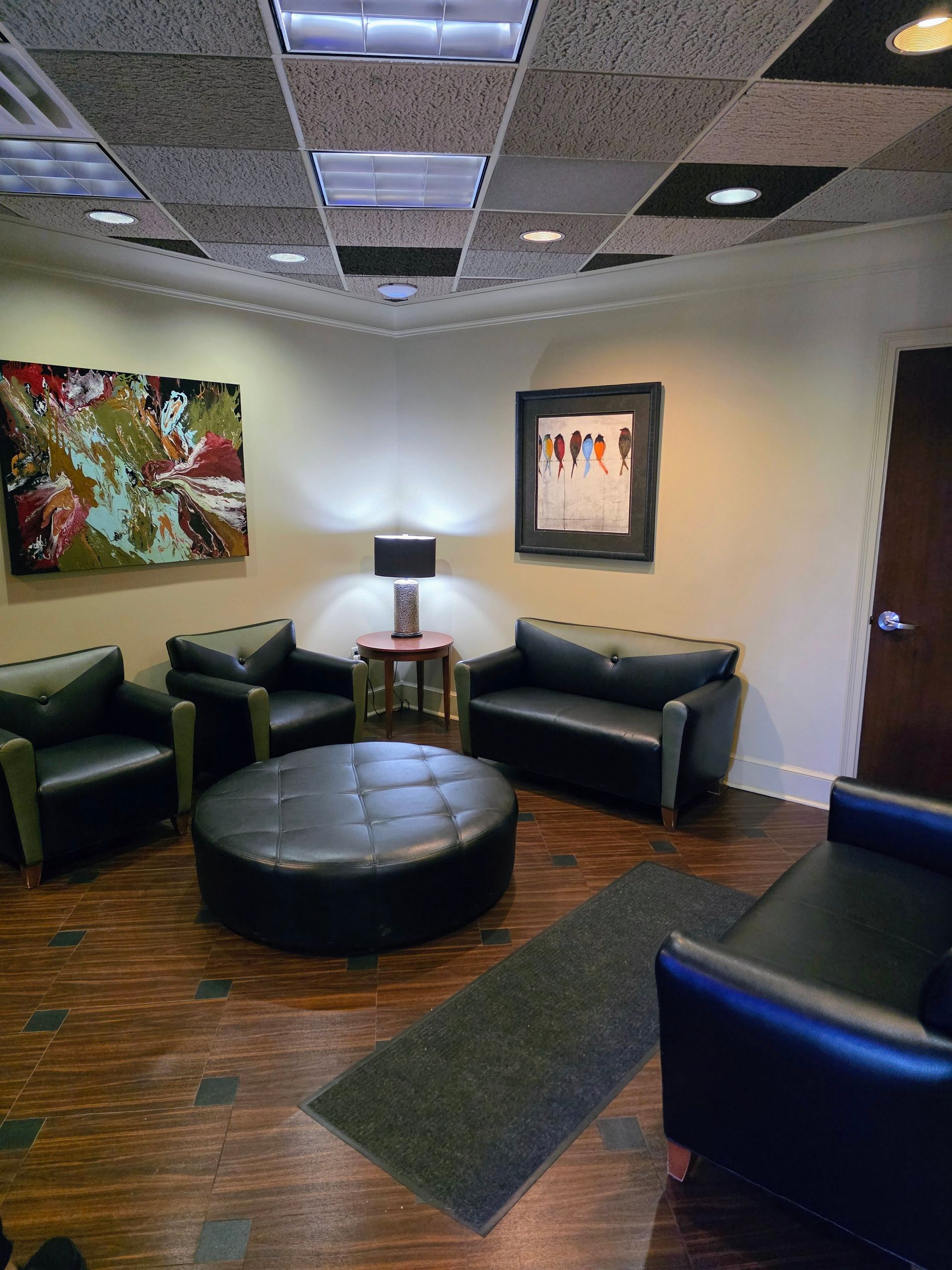 Waiting room with black leather furniture, abstract art, lamp, and dark wood-look flooring.