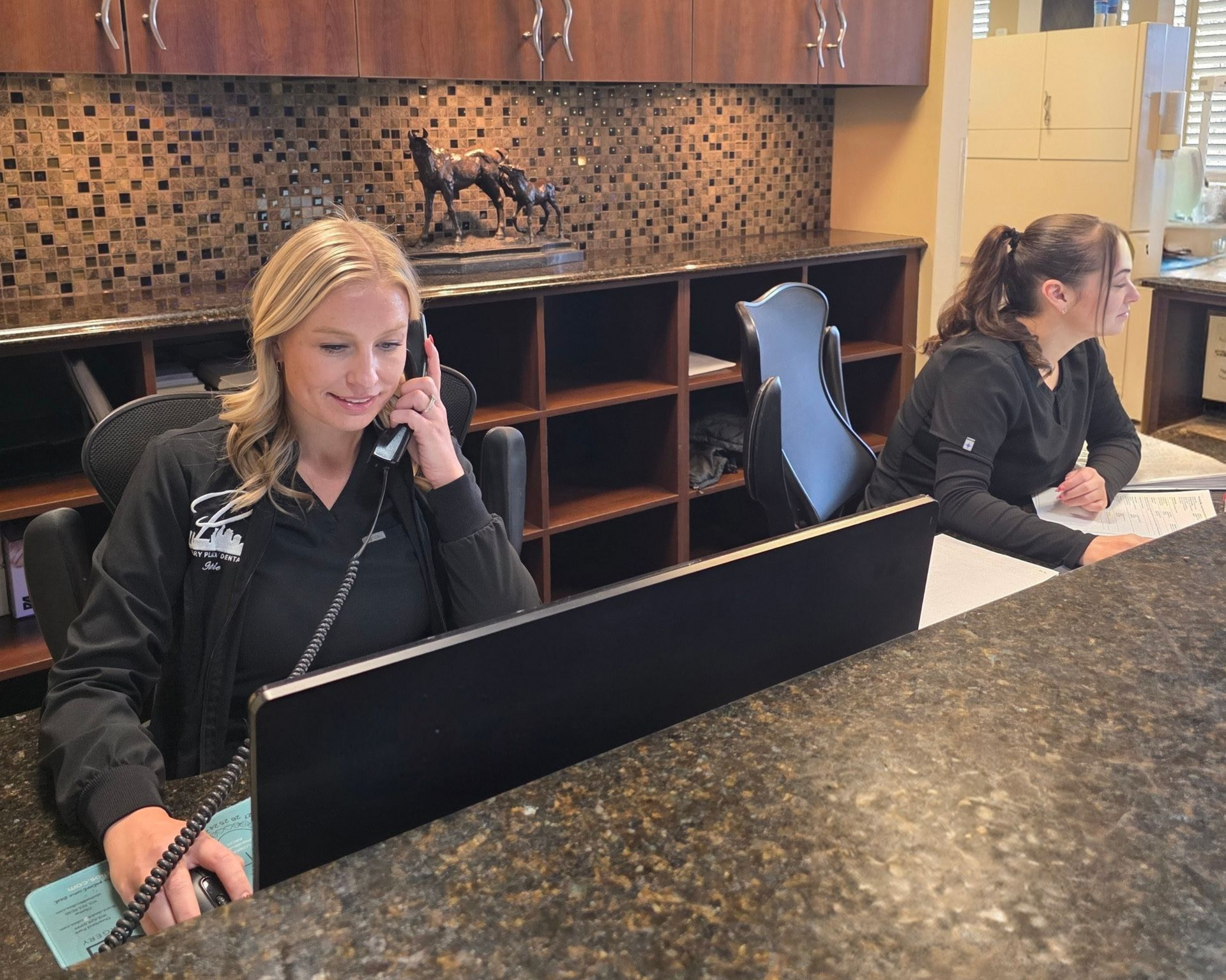 Two employees at a veterinary clinic reception desk. One answers phone, the other writes. Dark decor.