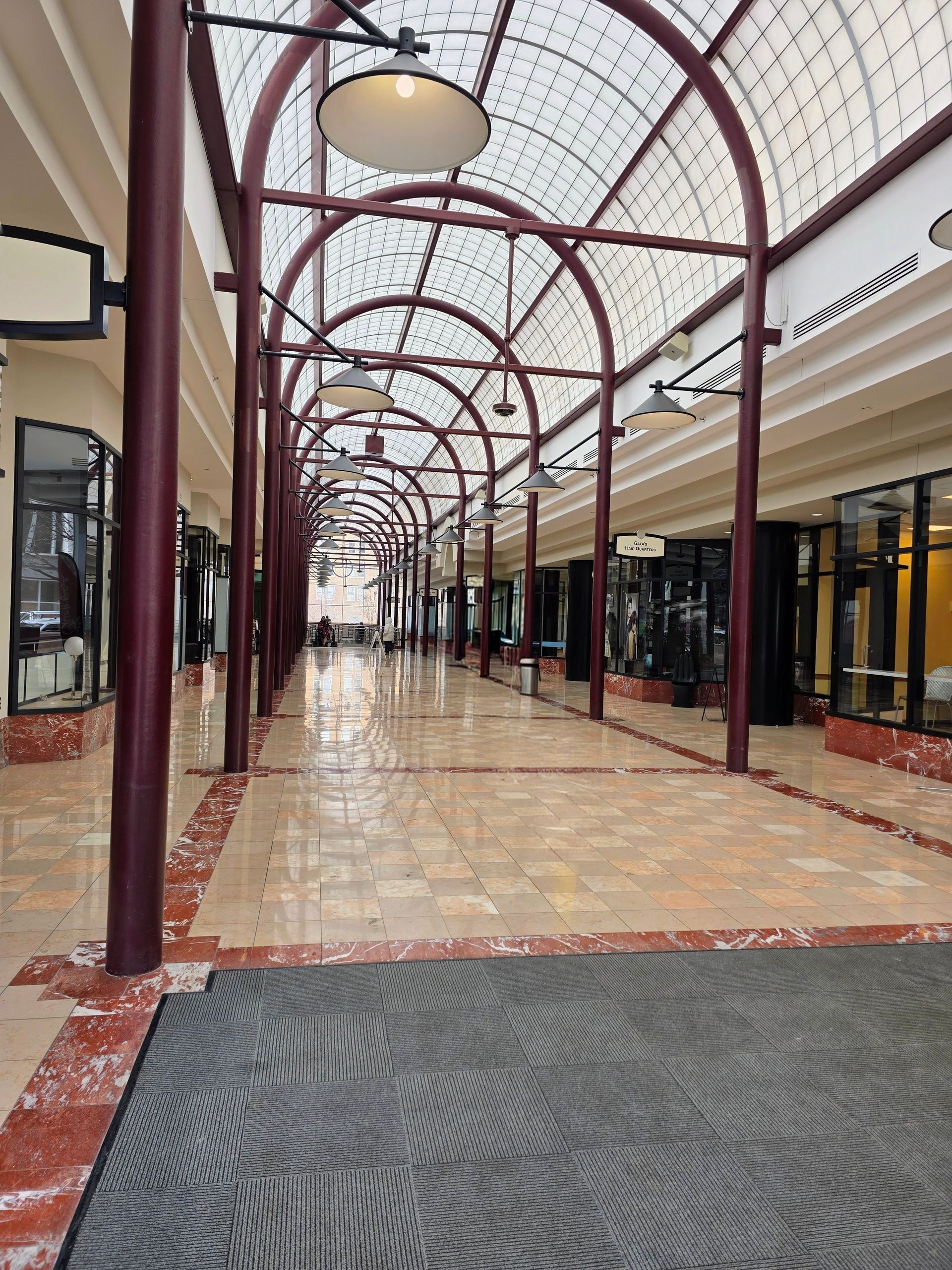 Interior view of a mall hallway with arched beams and shop fronts. Light-colored flooring and a patterned welcome mat.