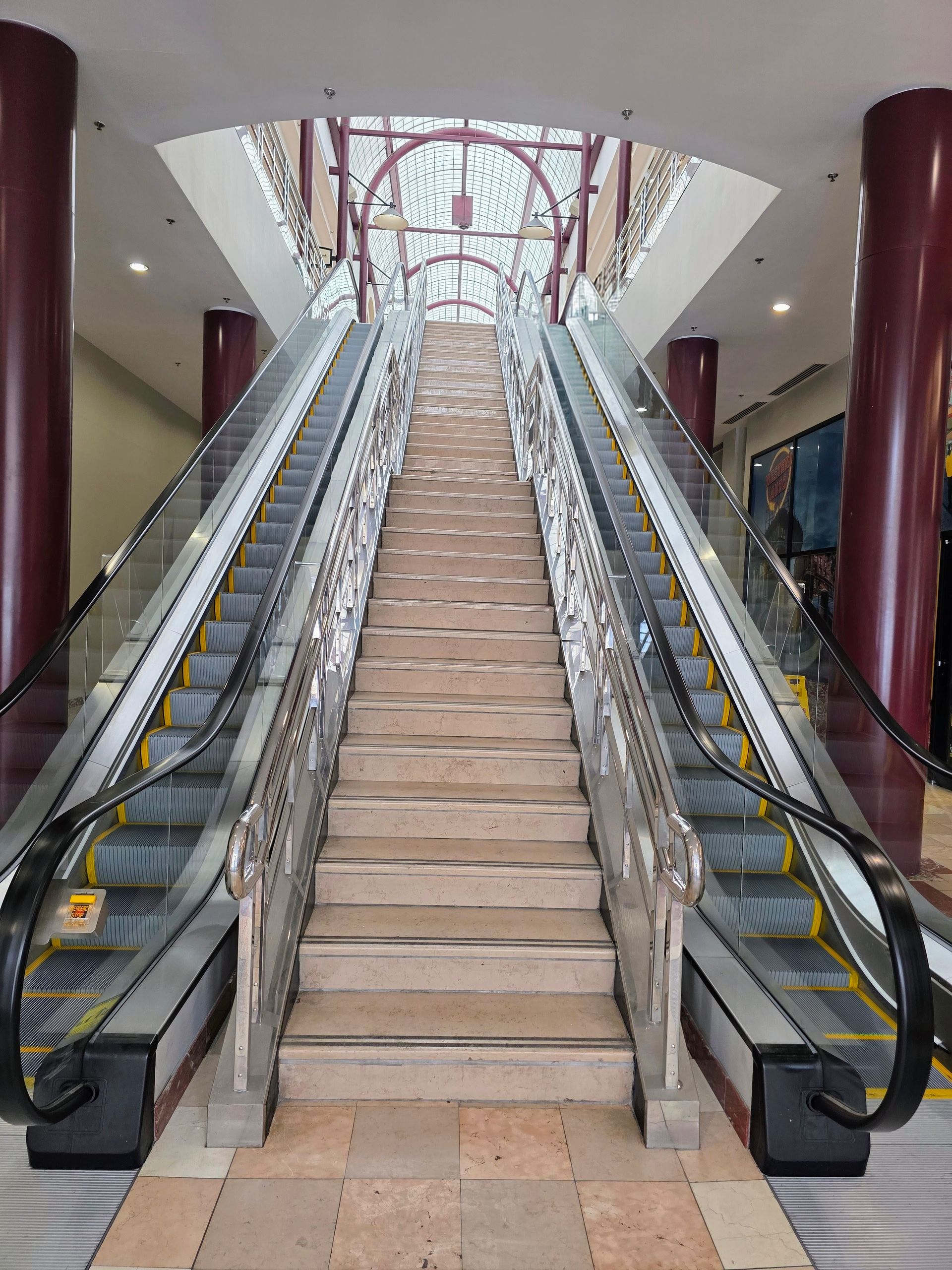 Escalators ascending in a commercial building. Red pillars, glass railings. Overhead decorative arch.
