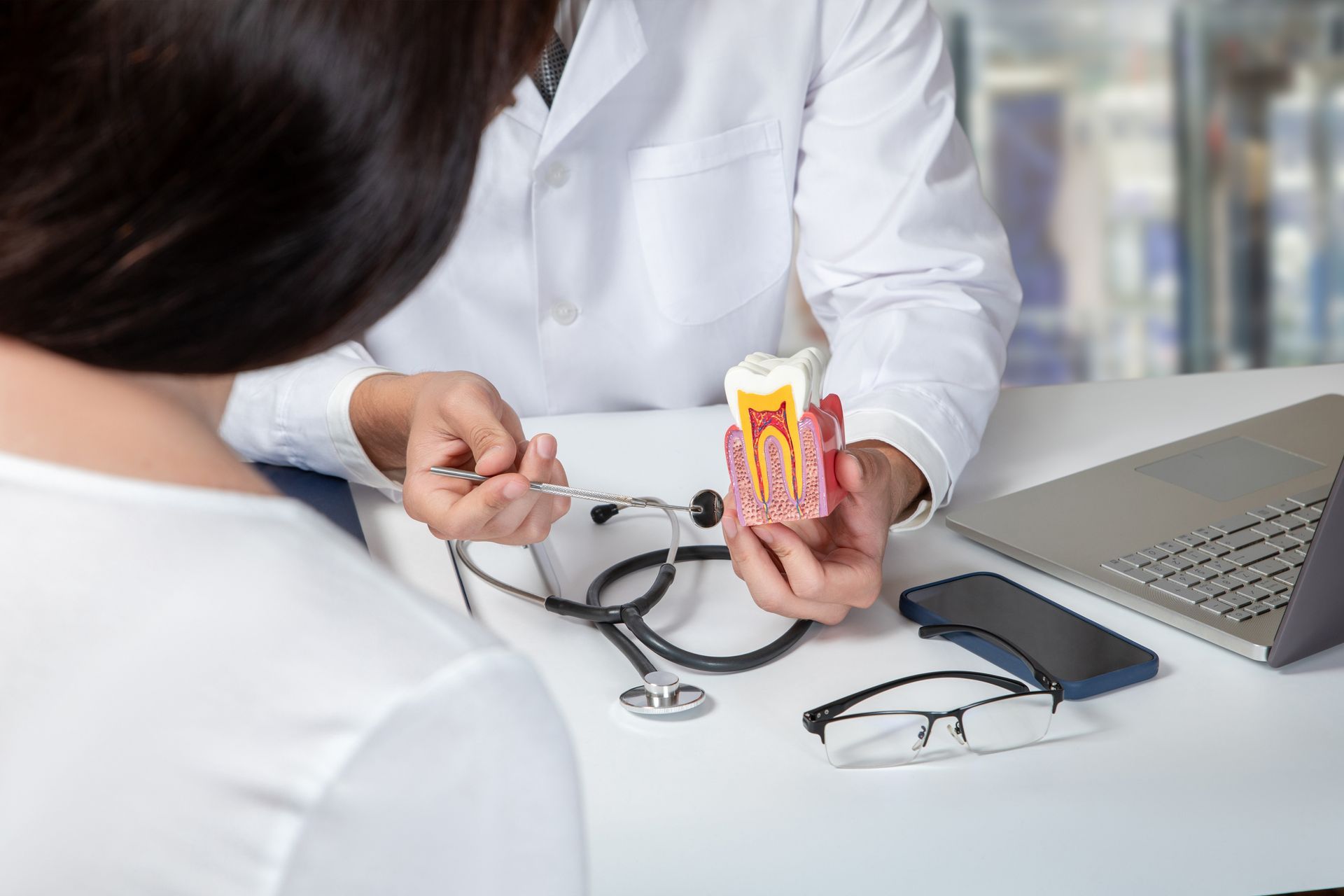 Dentist showing a tooth model to a patient in an office. Stethoscope, laptop, glasses on desk.