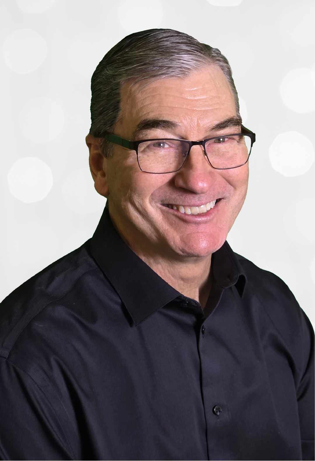 Man with glasses smiling, wearing a black shirt, against a blurred white backdrop.