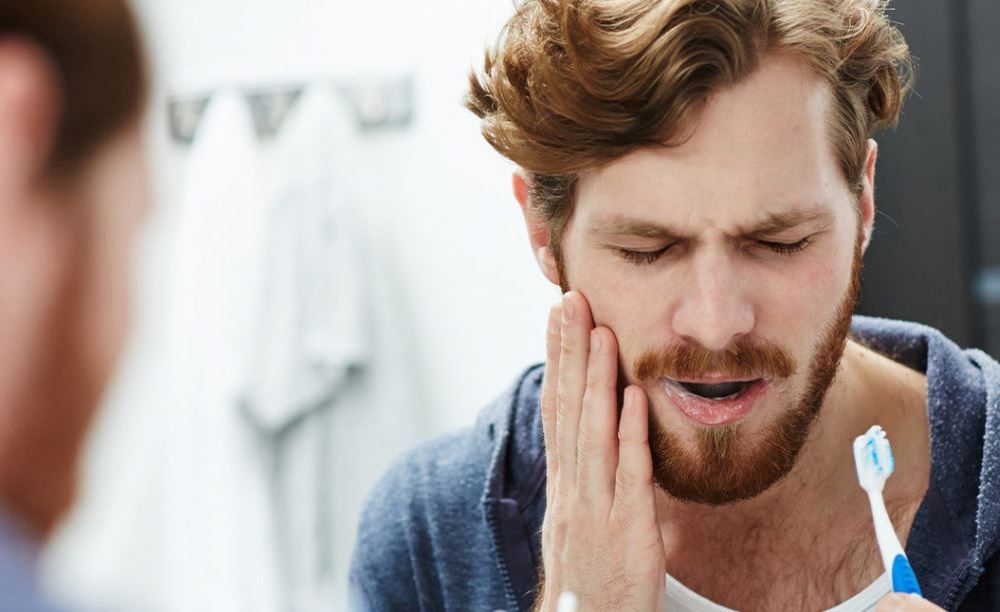 Man in bathroom with hand on cheek, grimacing, holding toothbrush, appearing to have a toothache.