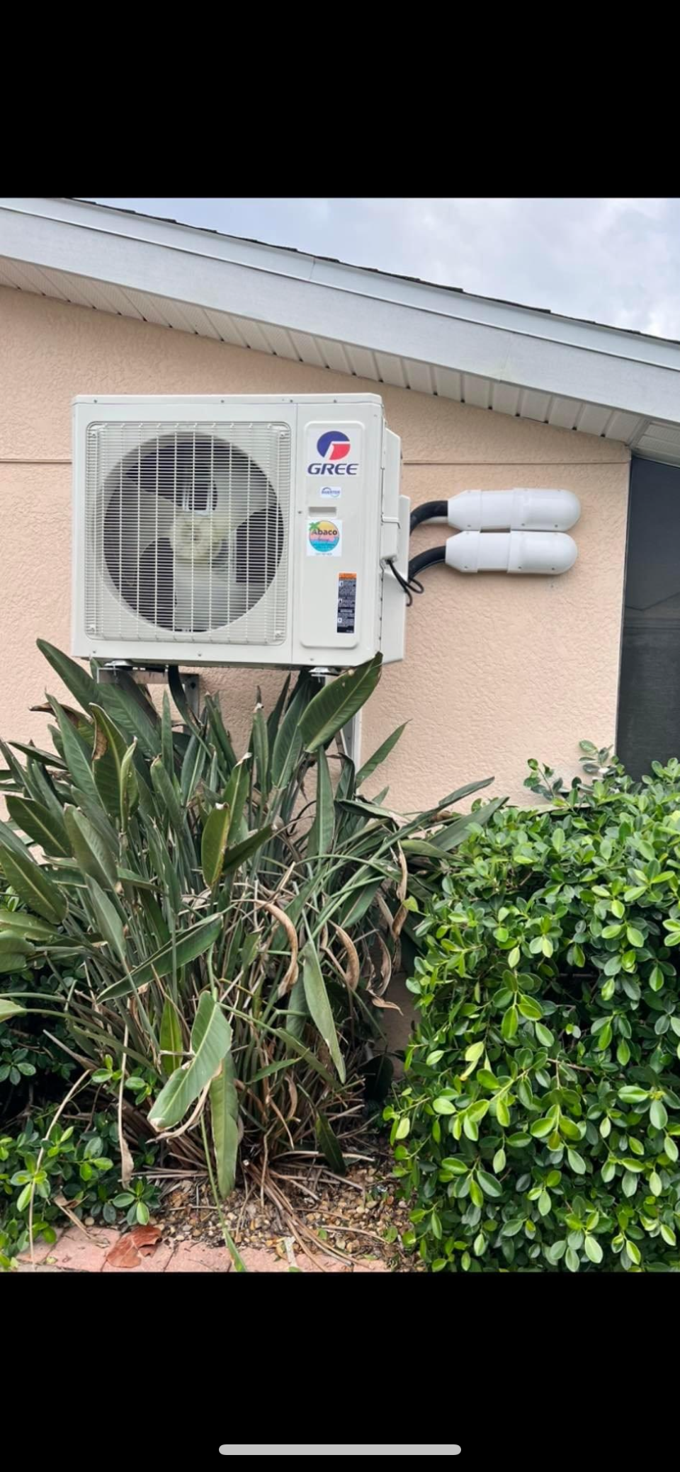 A white air conditioner is mounted on the side of a house next to a bush.