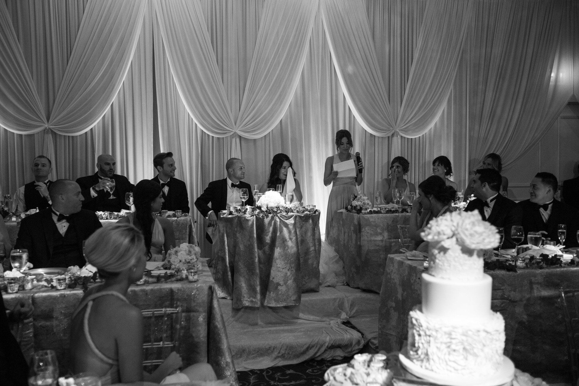 A black and white photo of a wedding reception with a cake in the foreground.