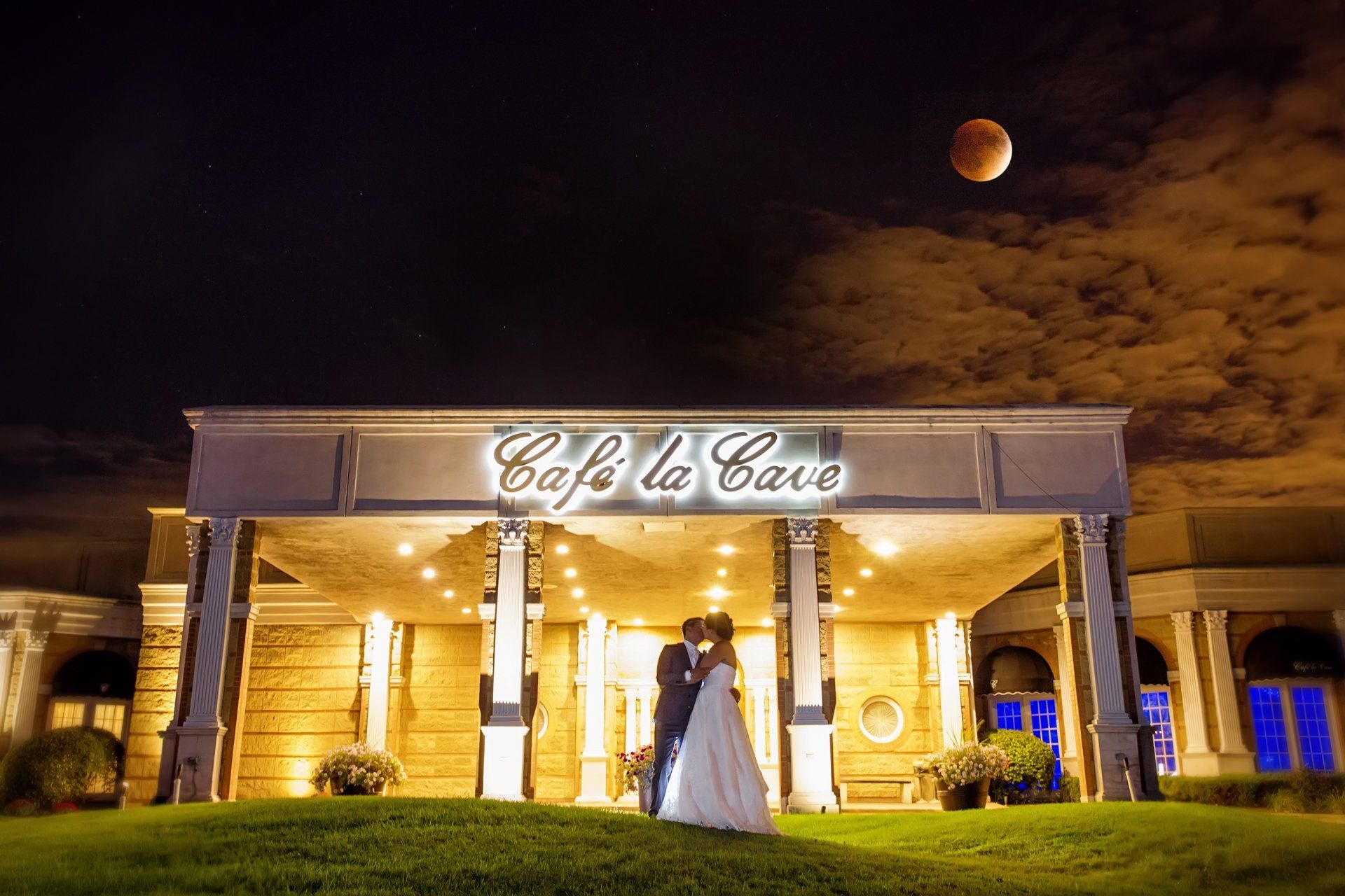 A bride and groom are kissing in front of a restaurant called cafe la cave