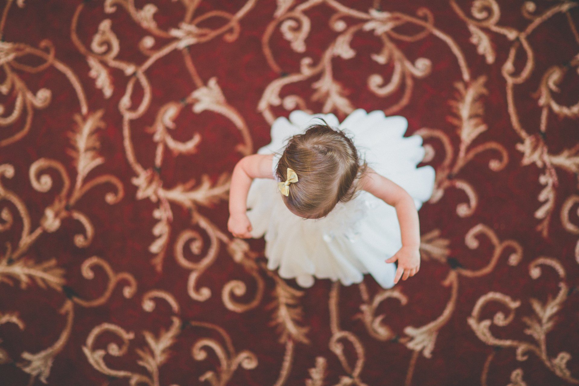 A little girl in a white dress is crawling on a red carpet.