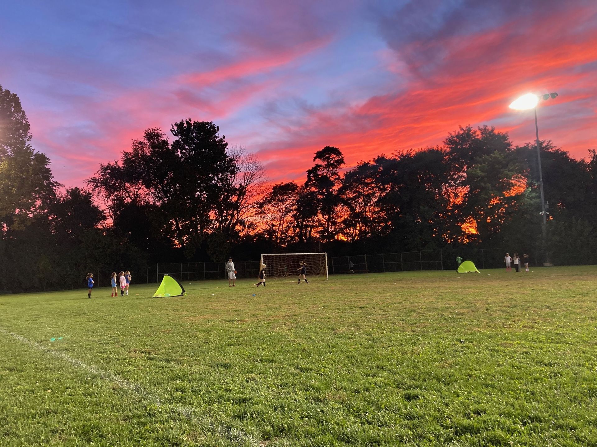 Sunset over a soccer field with a goal, and a few people under a pink-purple sky