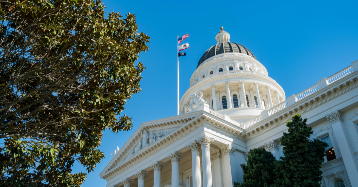 A low-angle shot of a grand neoclassical government building with a white dome and columns, set against a clear blue sky. A flagpole with the American, Californian, and another flag waves in the breeze. Green trees partially frame the structure, adding contrast to the bright architecture.