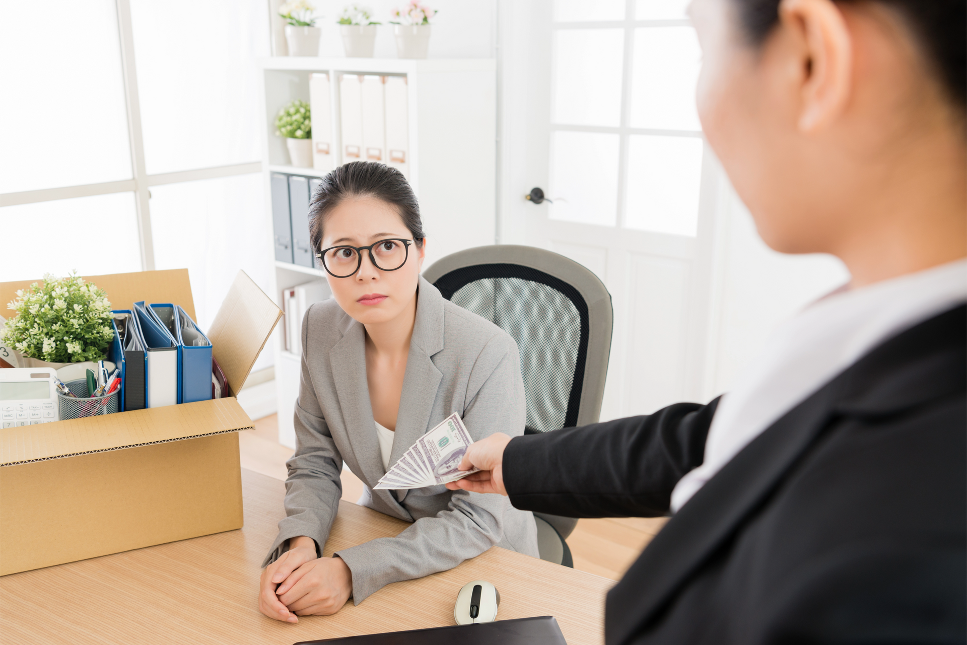 A woman is sitting at a desk with a box of her belongings and a woman is giving her money.