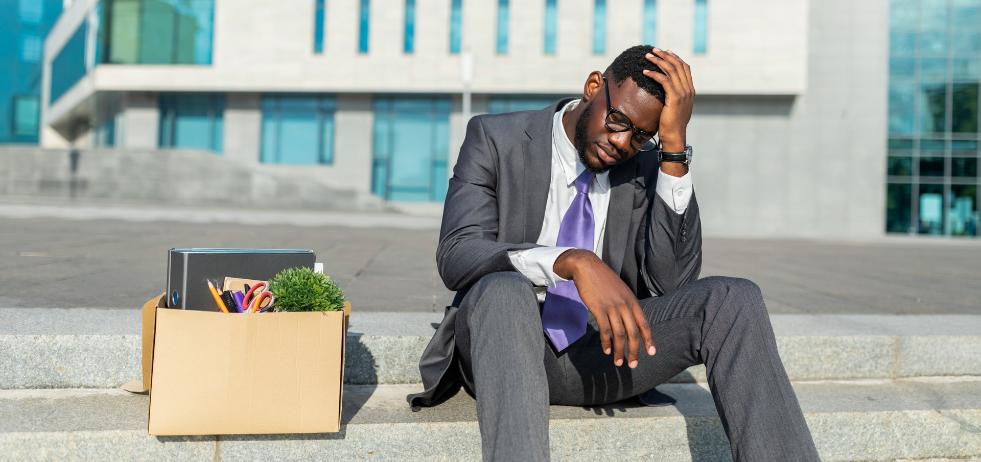 A professionally dressed man in a gray suit and purple tie sits on outdoor concrete steps, looking distressed with his head resting on one hand. A cardboard box filled with office supplies, including a binder, scissors, and a small plant, sits beside him, suggesting he has been laid off or left his job. The background features a modern office building with large windows.