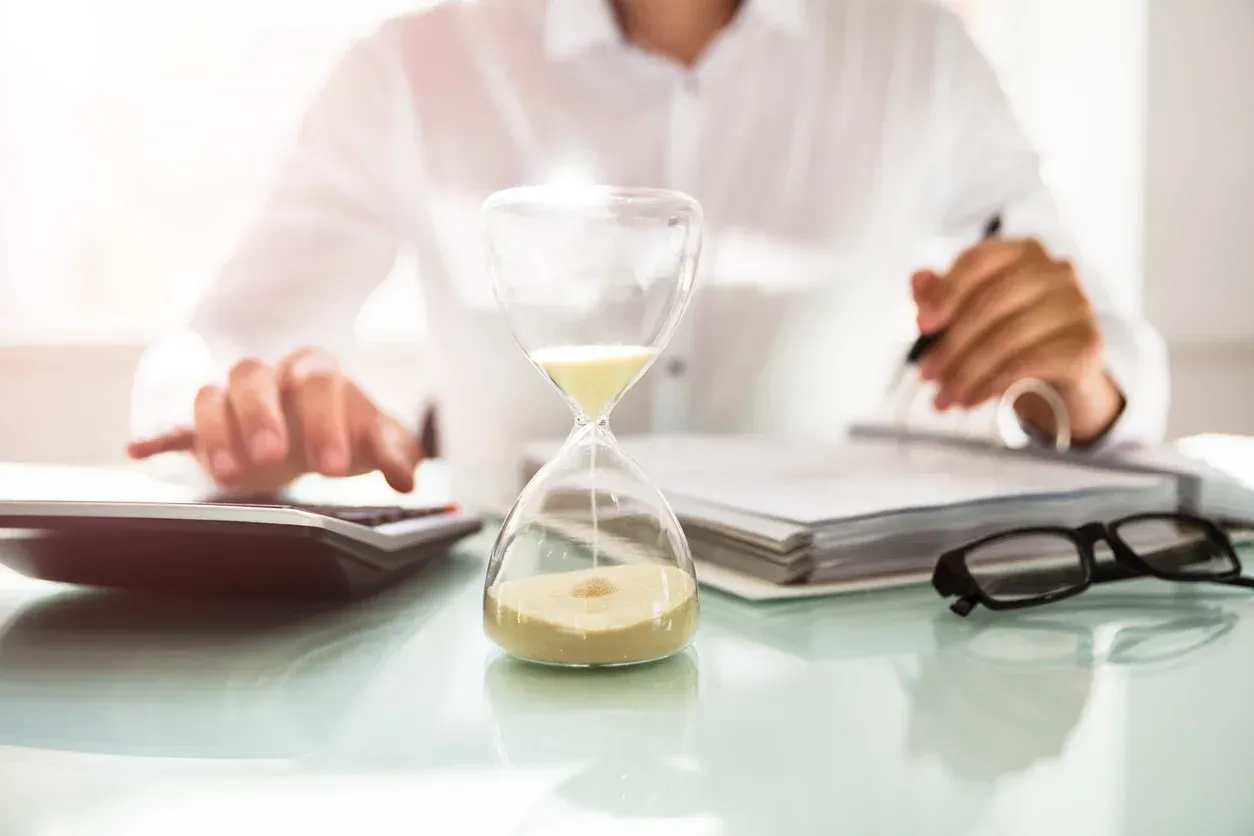 Person using a calculator and writing, with an hourglass and glasses on a desk.