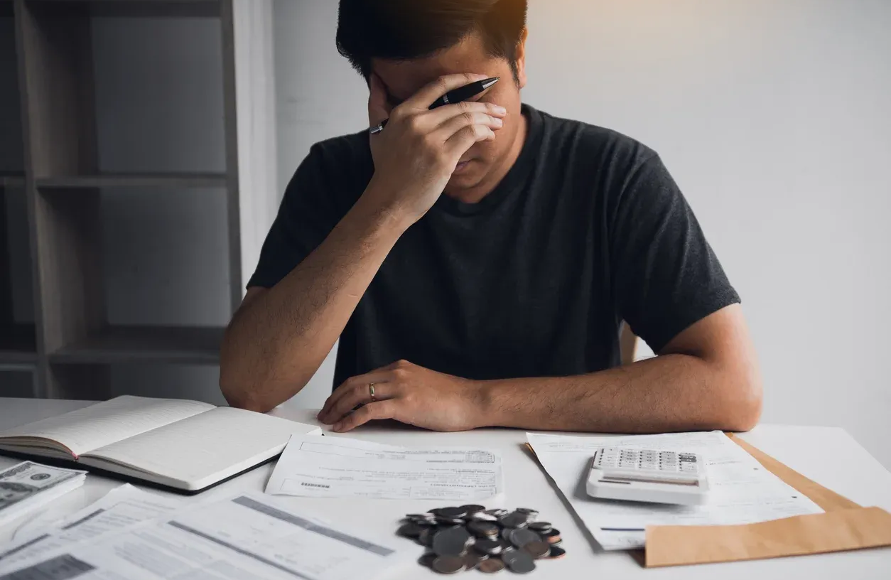 Man at a desk with head in hand, stressed, looking at bills, and loose change.