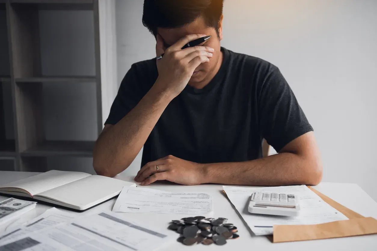 Man at a desk with head in hand, stressed, looking at bills, and loose change.