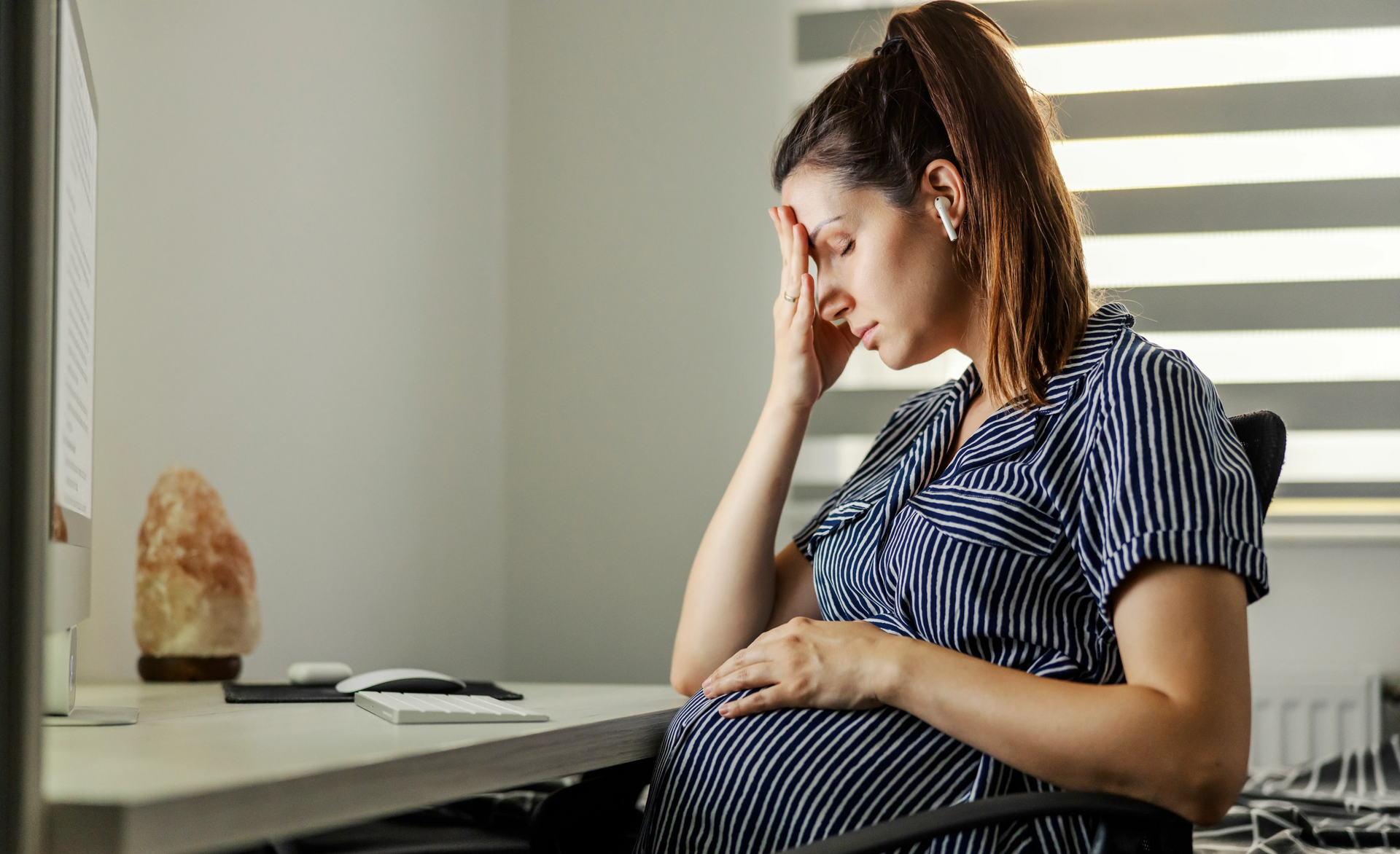Pregnant woman with hand on forehead, seated at a desk, looking stressed, indoors.