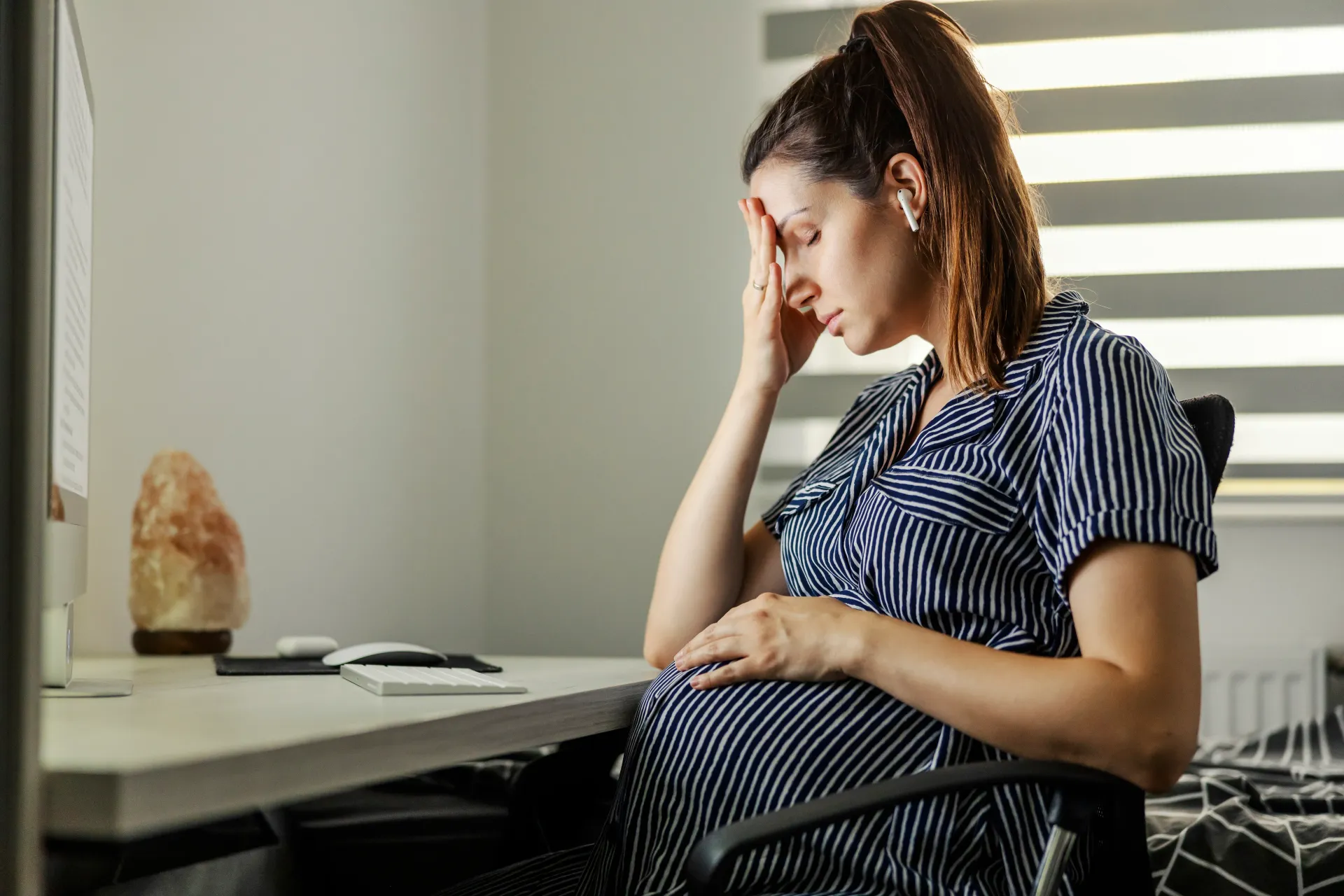 A pregnant woman is sitting at a desk with her hand on her forehead.