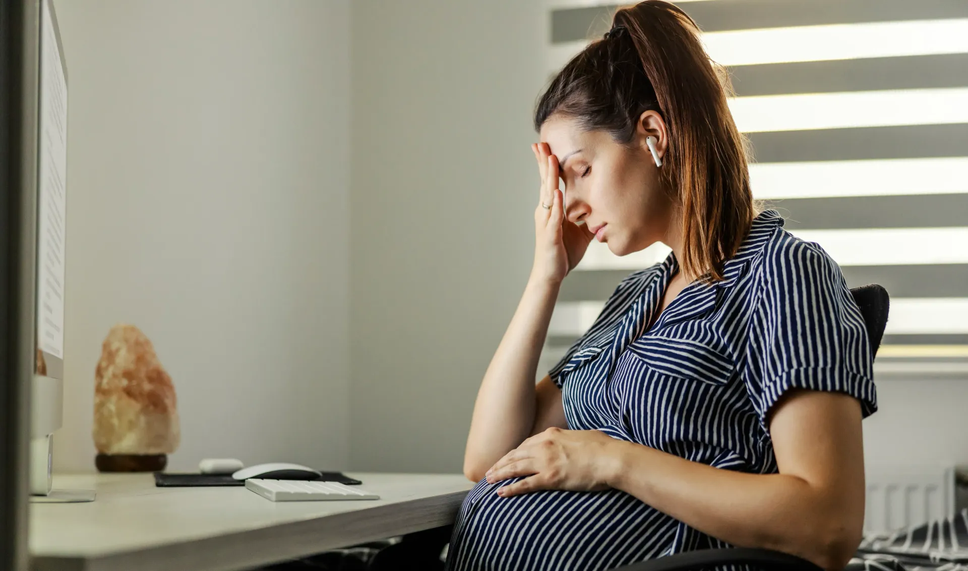 A pregnant woman is sitting at a work desk with her hand on her forehead, visibly distressed.