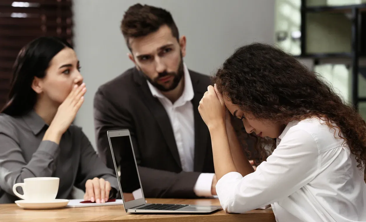 Woman with head in hands, sitting at a table with a laptop, while two coworkers whisper, looking concerned.
