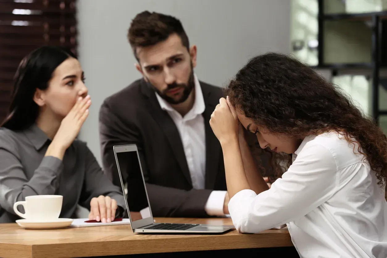 Woman with head in hands, sitting at a table with a laptop, while two coworkers whisper